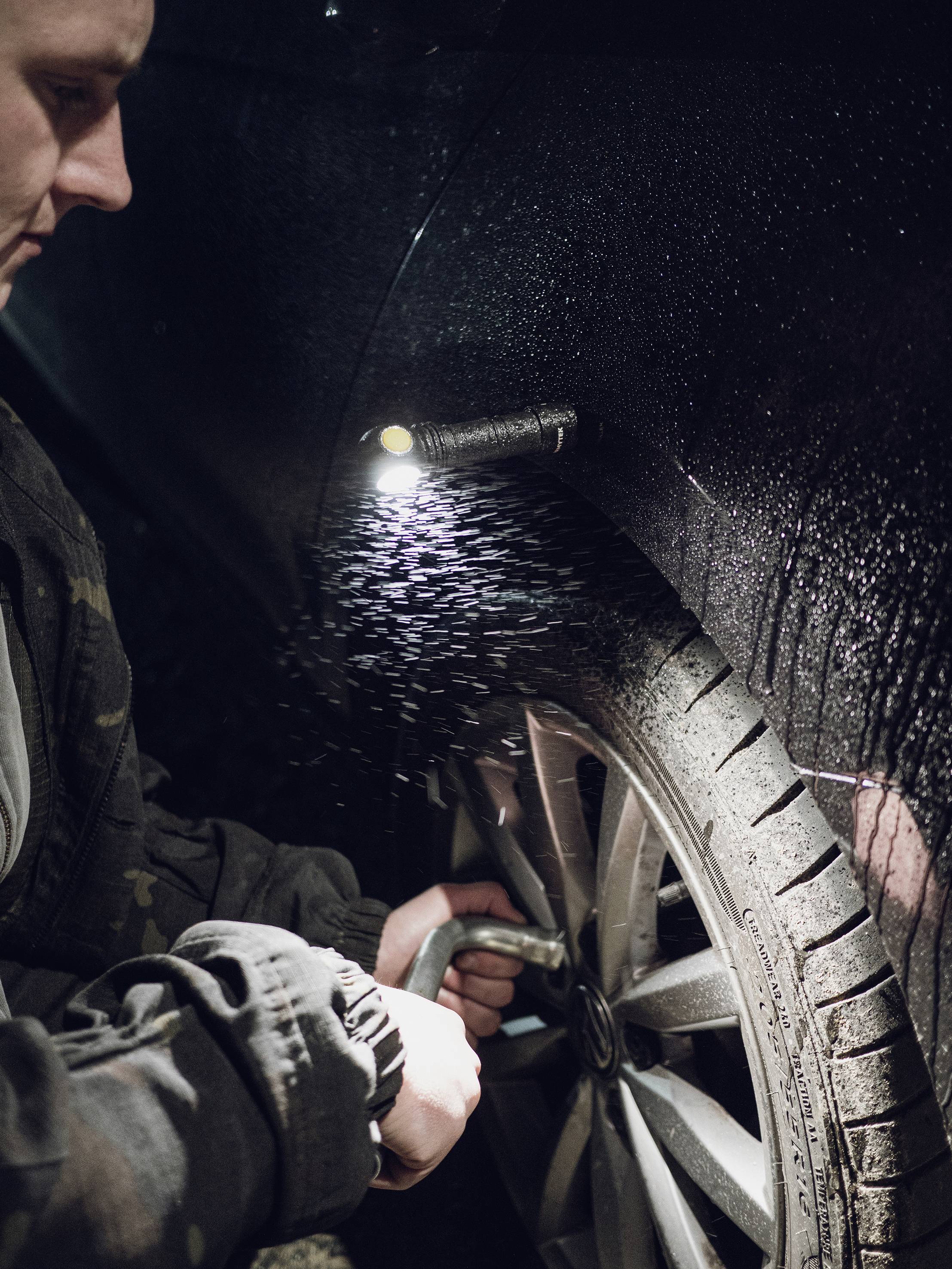 A person is checking the tyre pressure of a car at night, illuminated by a small lamp.