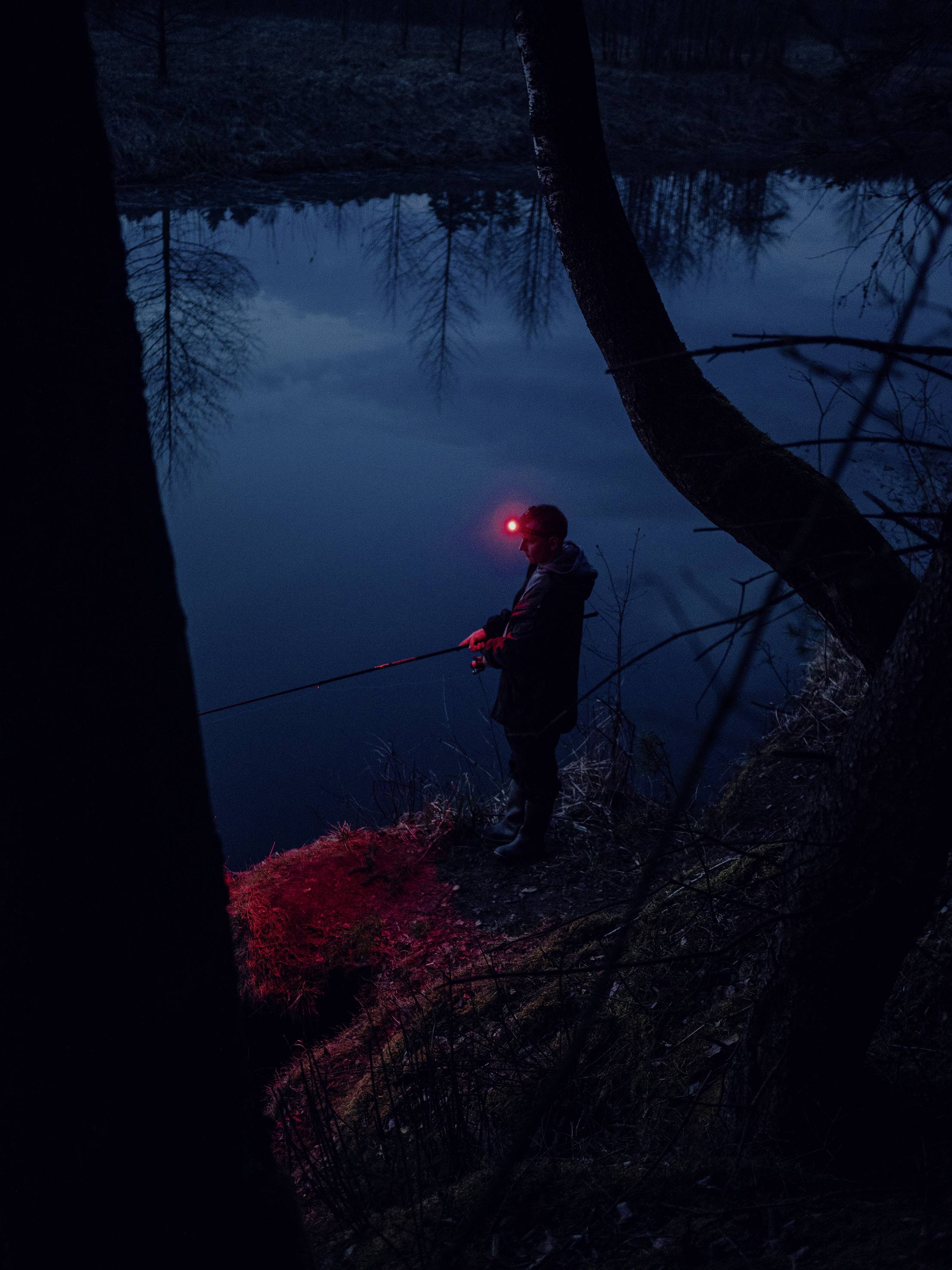 A person wearing a head torch fishes at night along a riverbank.