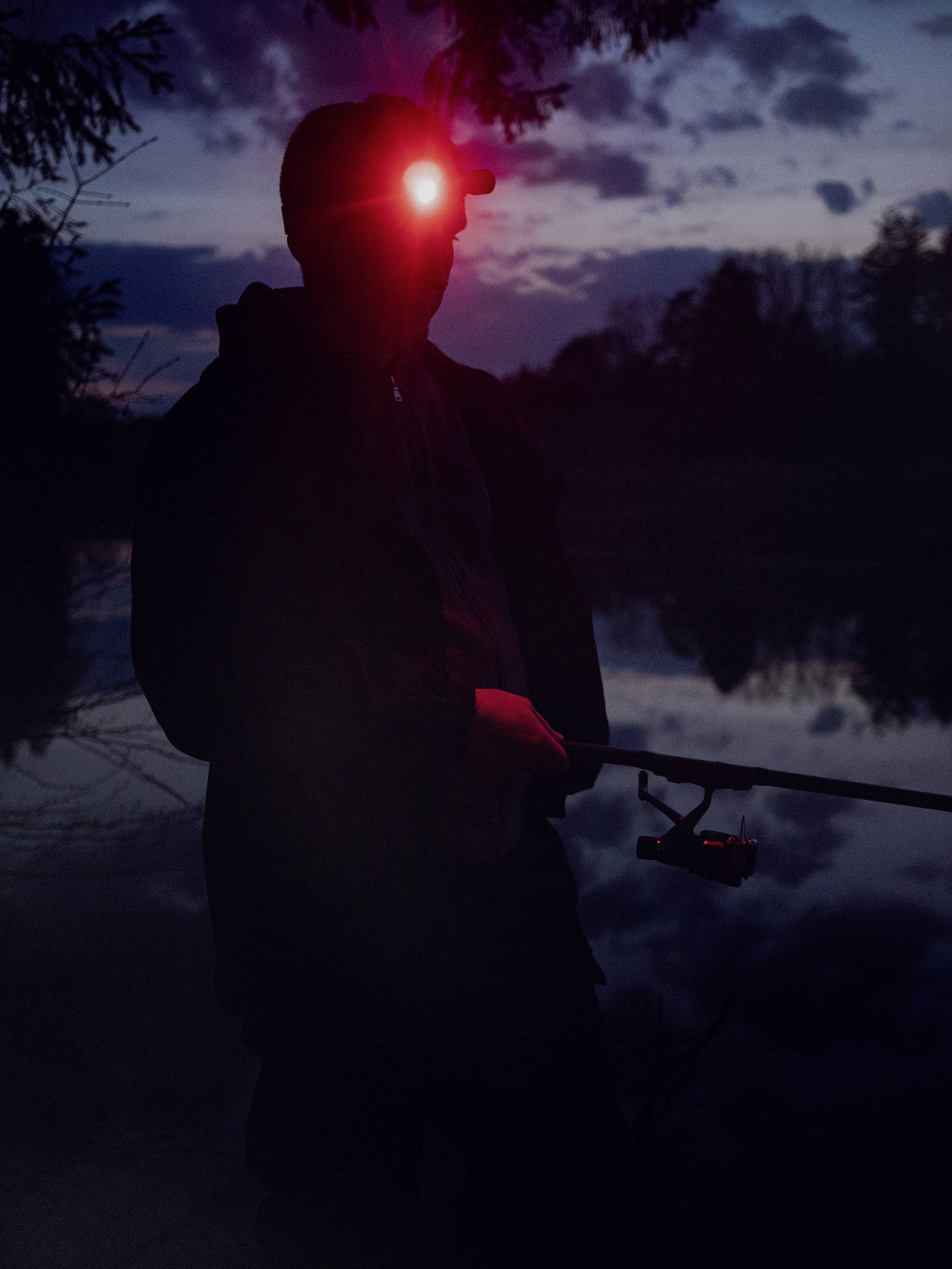 A person stands fishing by the tranquil lakeside at night, a red head torch illuminating the darkness.