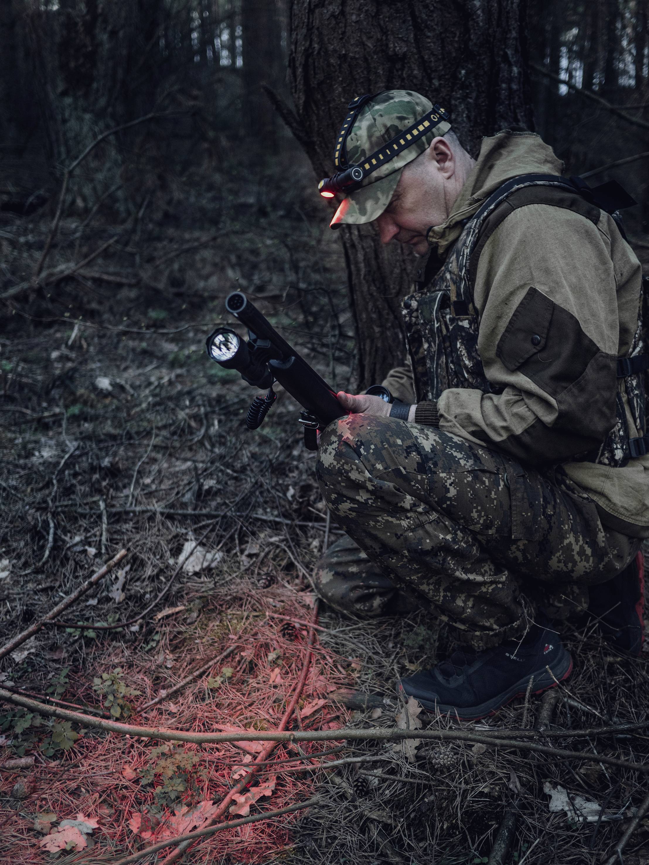 A hunter in camouflage crouches in the woods, examining the ground with a torch. Red light illuminates the foliage.