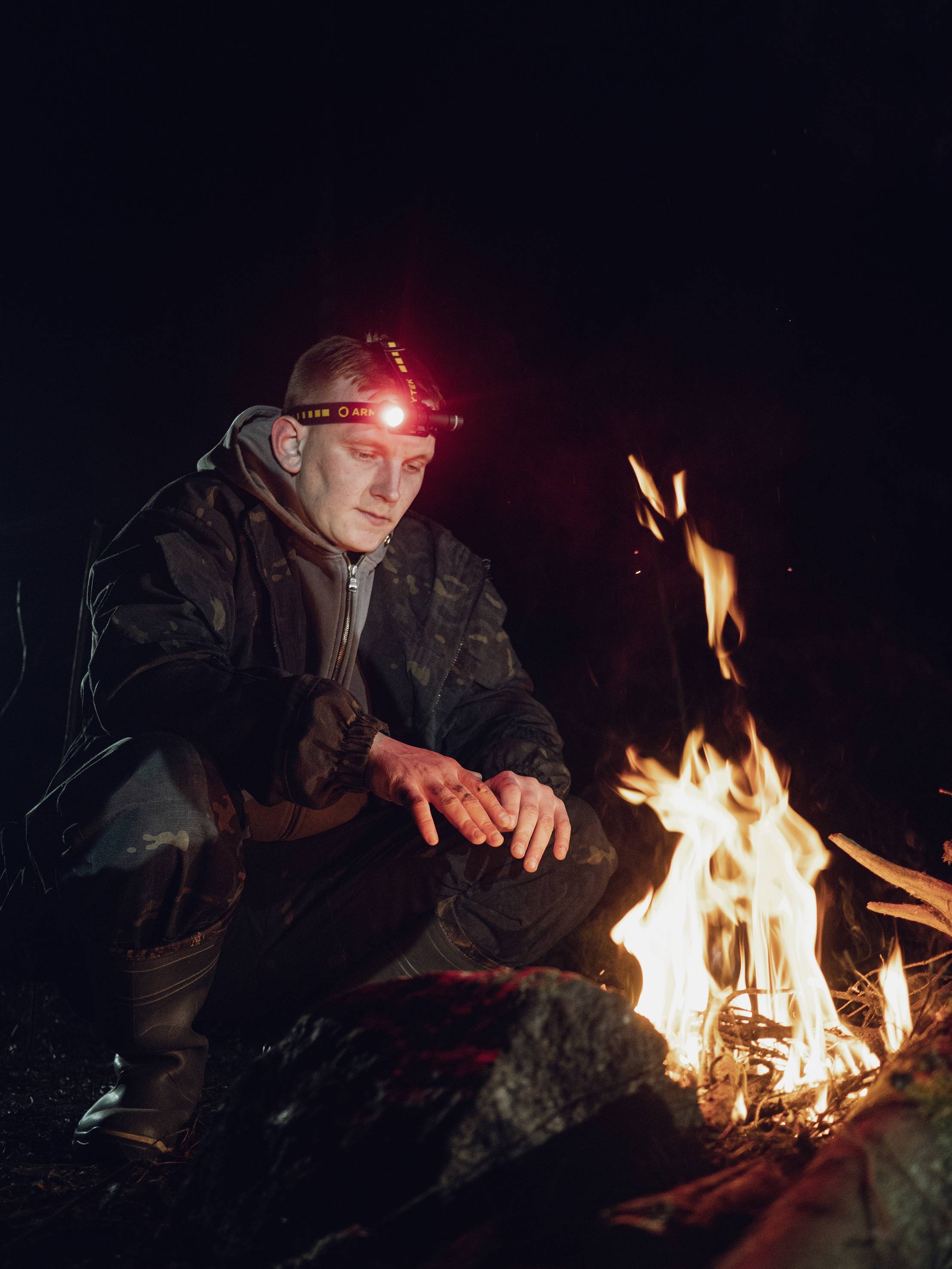 A person wearing a head torch sits outdoors at night by the campfire. They are warming their hands by the flames.