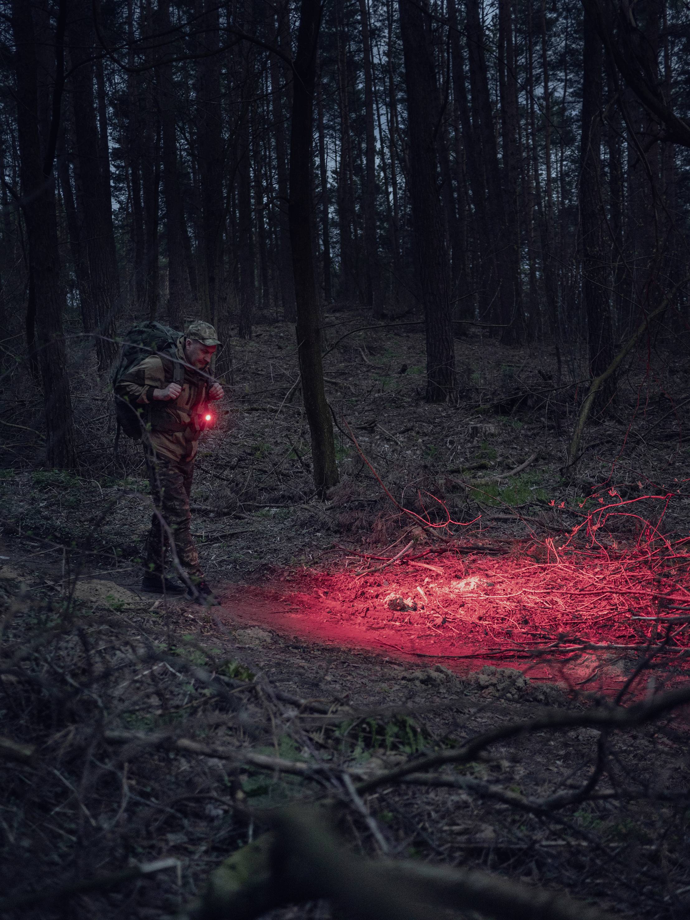 A person in camouflage stands in the woods at night, illuminating a red glow on the ground. The surroundings are dark and wooded.