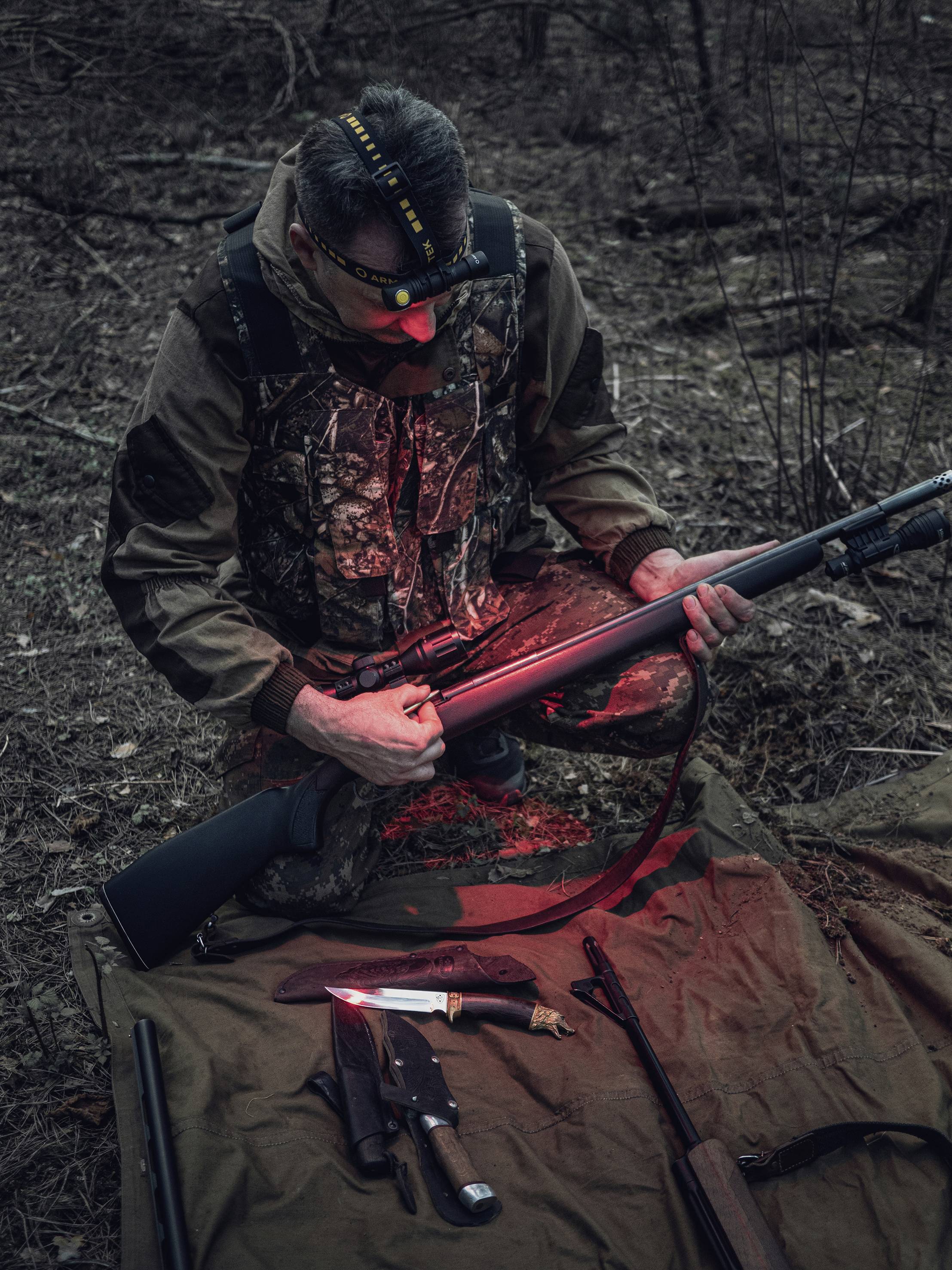 The hunter kneels in the woods and examines a rifle in dim light. A knife and tools are laid out on a tarpaulin.