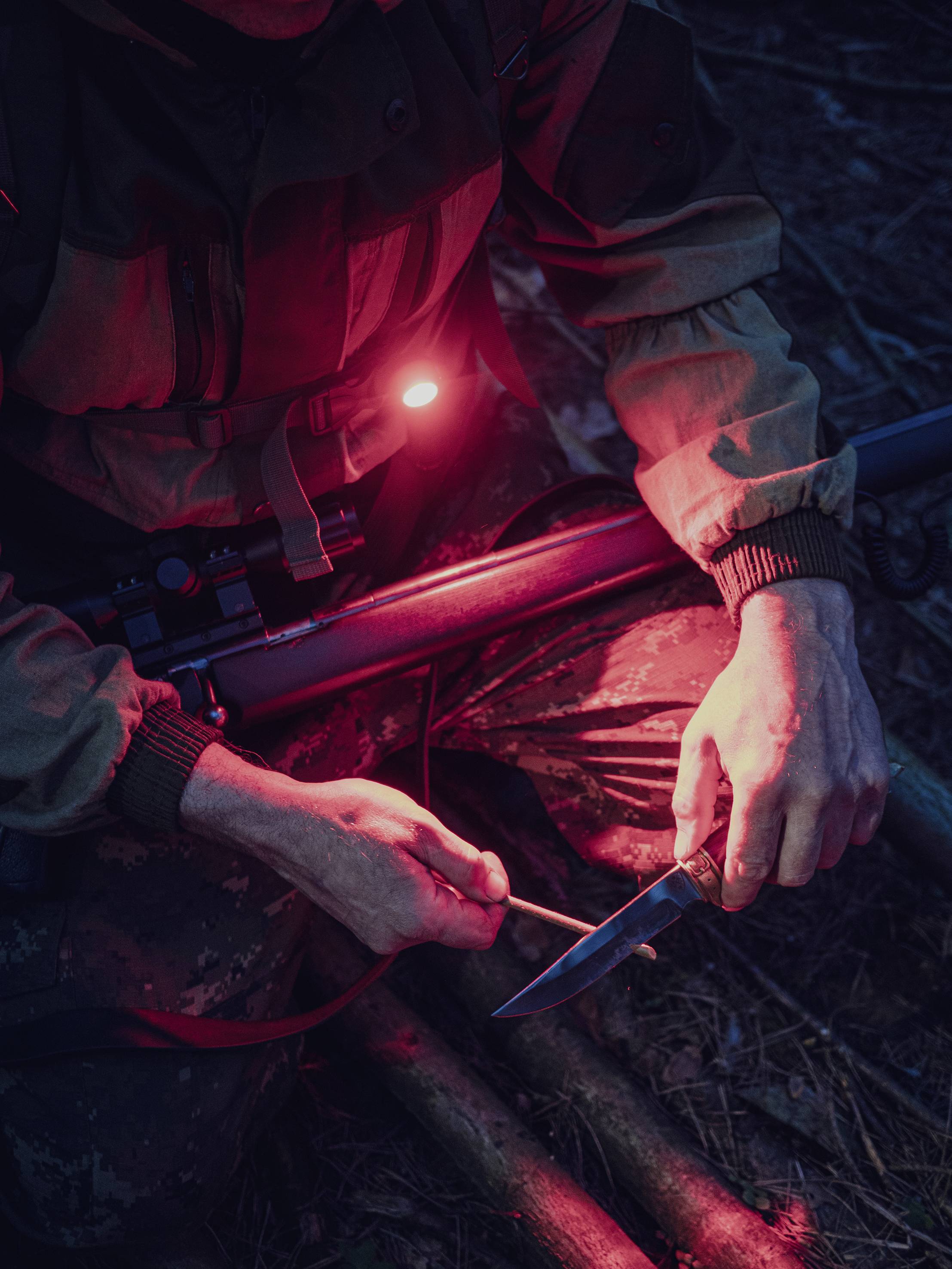 A person sits in darkness in the woods, holding a knife, illuminated by a red light. They are wearing camouflage clothing.