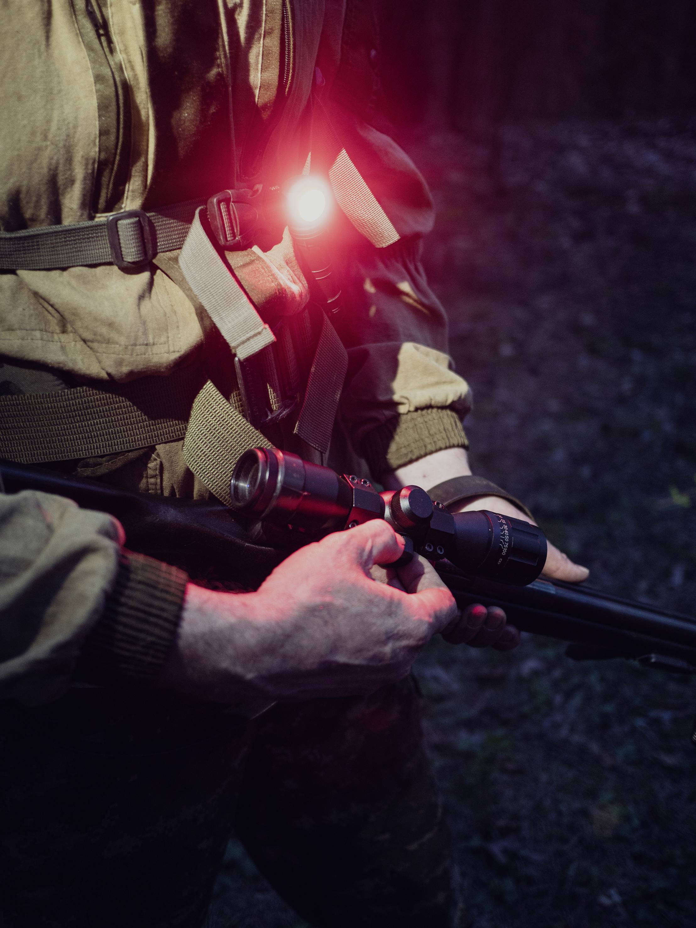 A person in camouflage clothing is holding a rifle with a telescopic sight. A red light is glowing from the chest pocket.