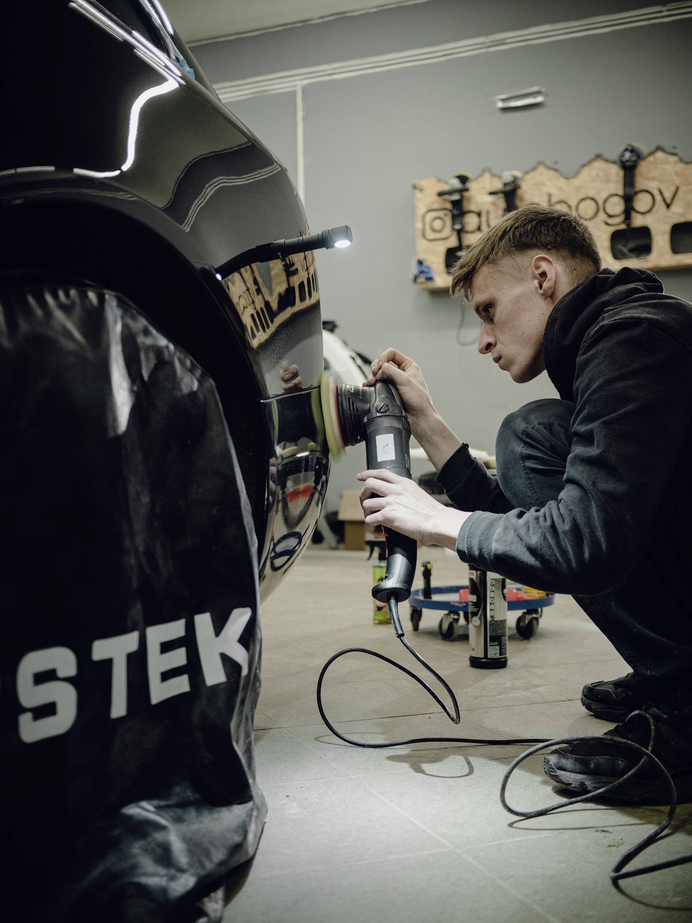 A young man is polishing a black car in a workshop. A tool wall can be seen in the background.