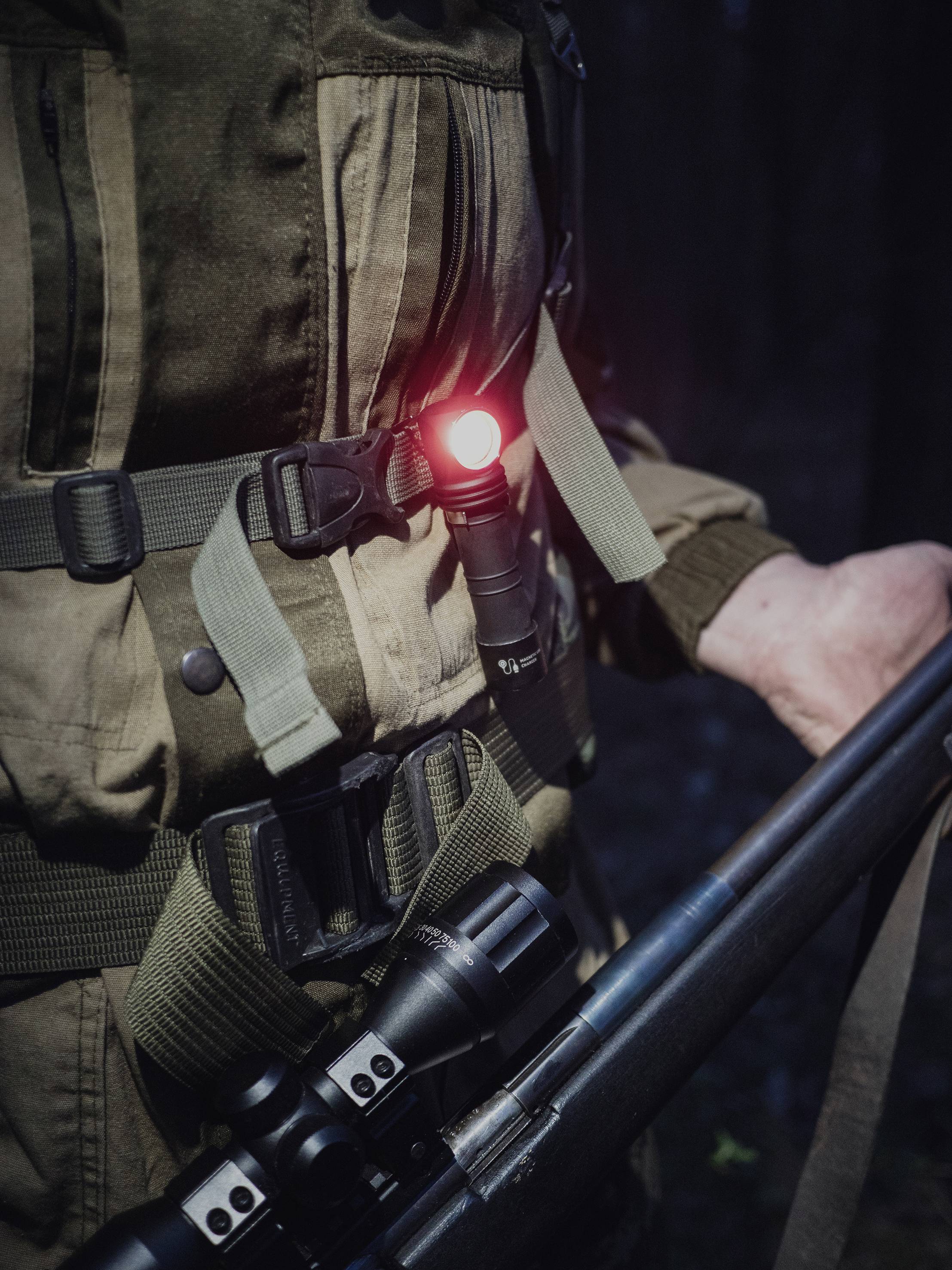 A person in dark clothing holds a rifle in the woods, with red light on their equipment.