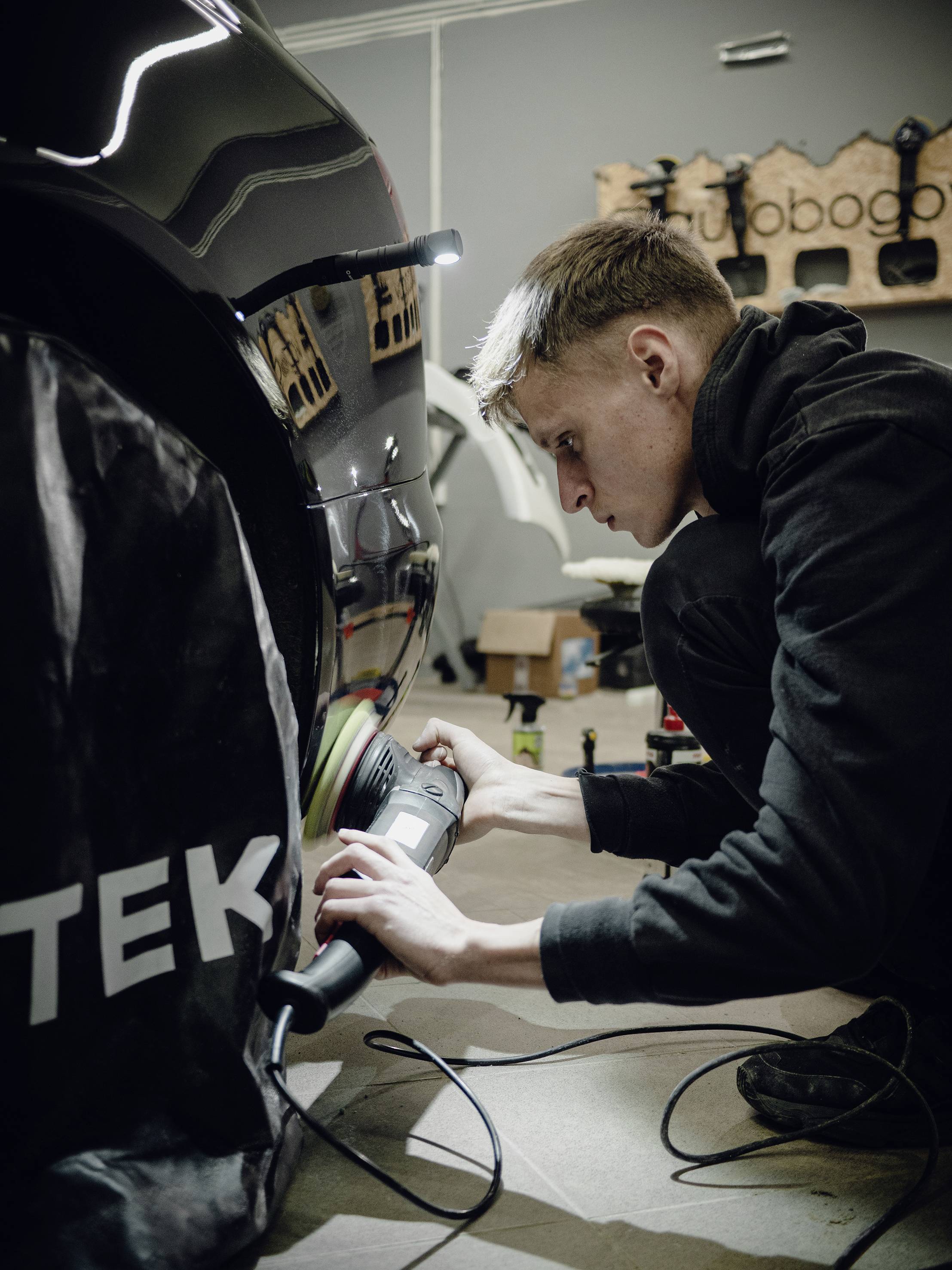 A man is polishing a black car in a workshop. He is focusing intently on his work. Tools are visible in the background.