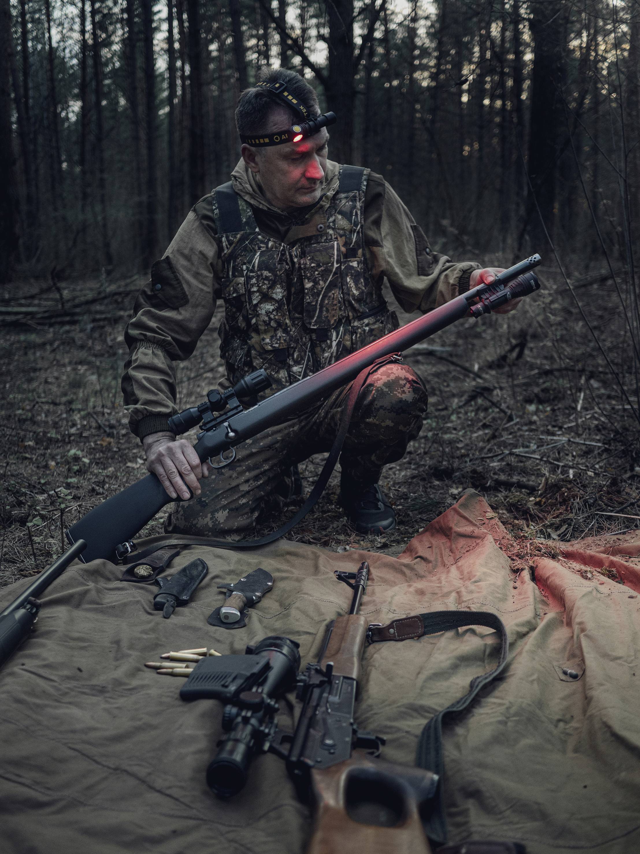 A hunter sits on one knee in the woods, holding a rifle. Two rifles and ammunition lie on a tarpaulin in front of him. Twilight is visible in the background.