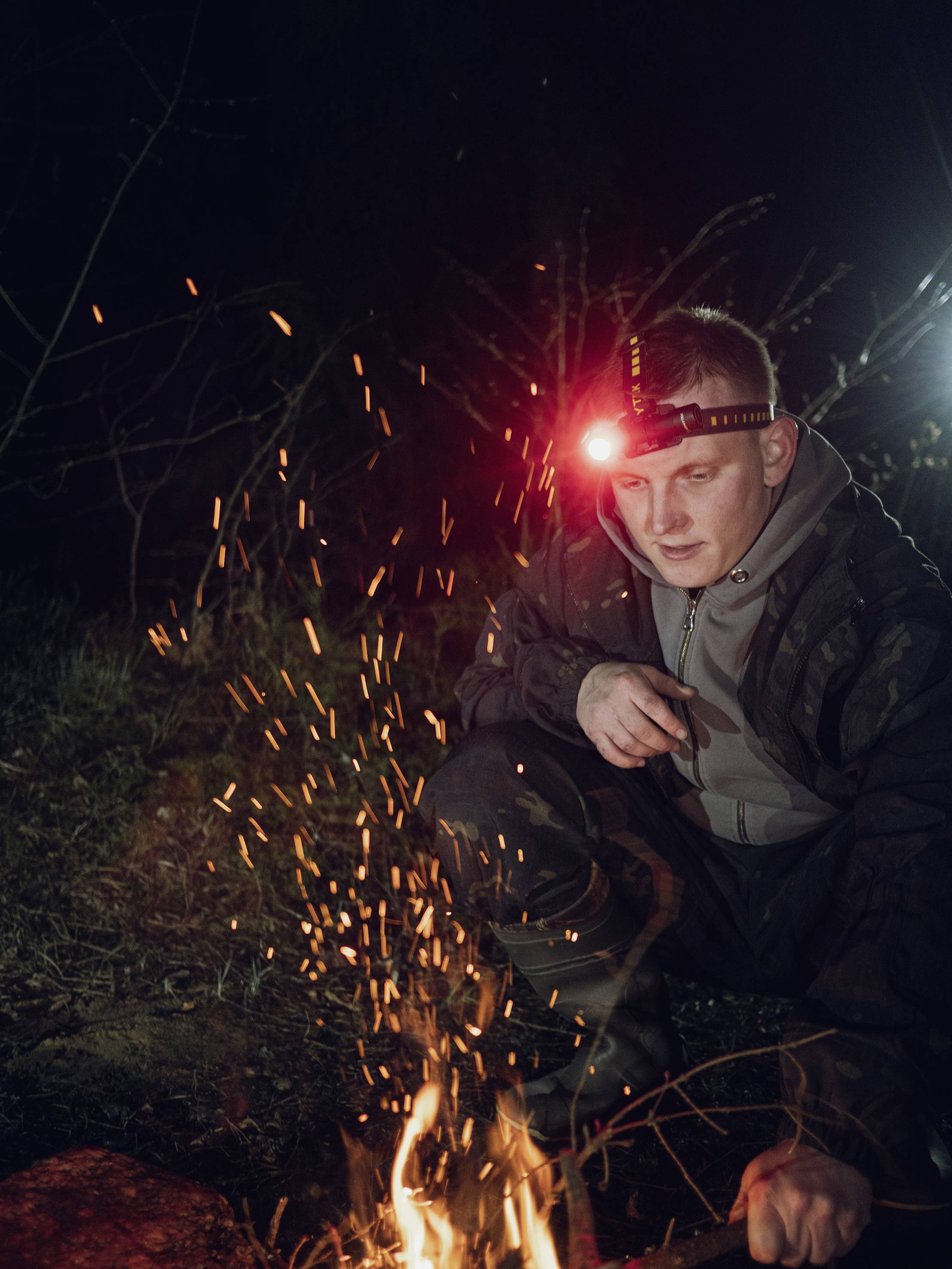 A person wearing a head torch kneels in the darkness and observes a small campfire, with sparks flying from it.