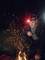 A person wearing a head torch kneels in the darkness and observes a small campfire, with sparks flying from it.