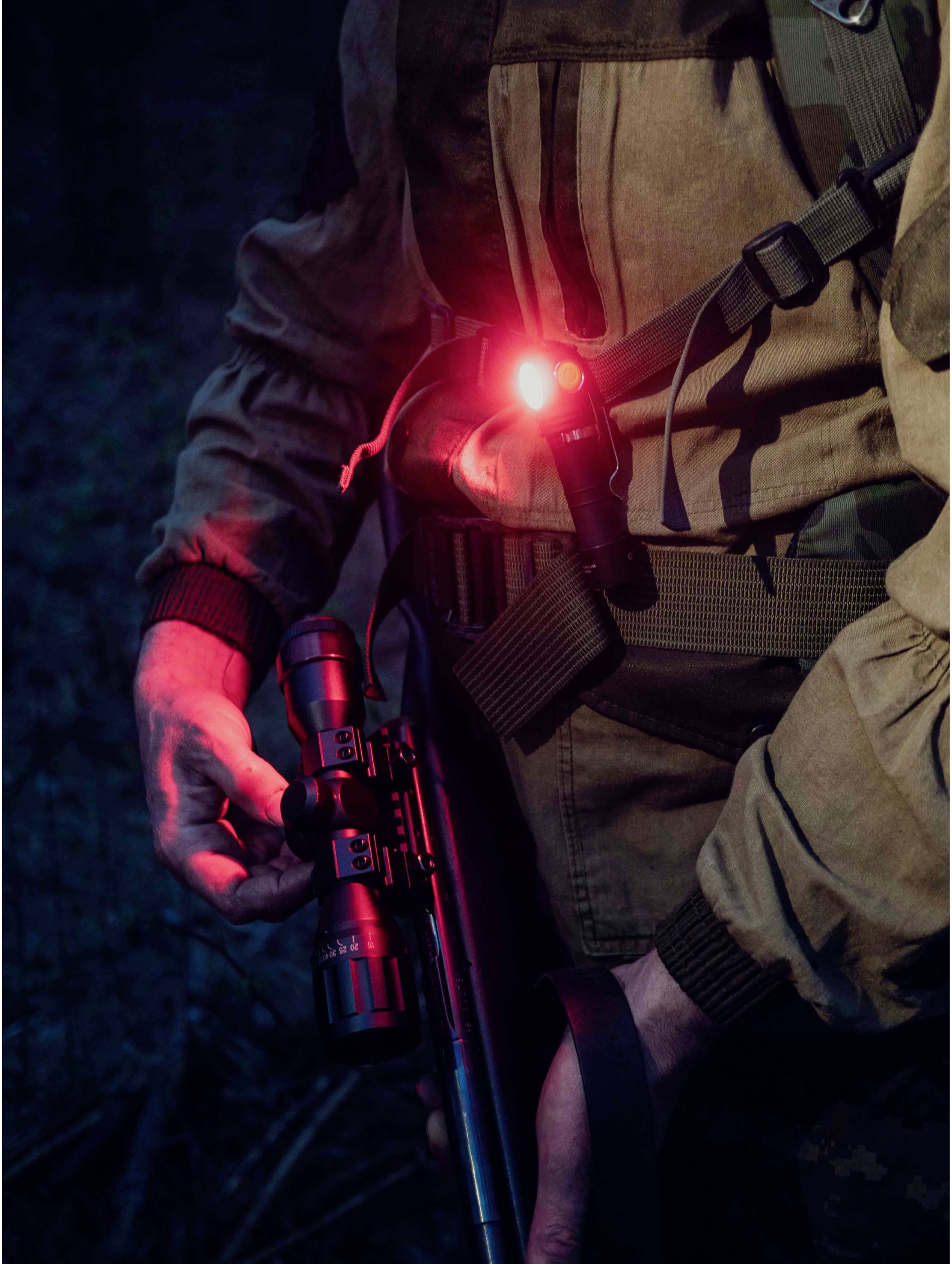 A person in a dark forest is carrying a rifle and a red light on their left arm, which slightly illuminates the surrounding area.