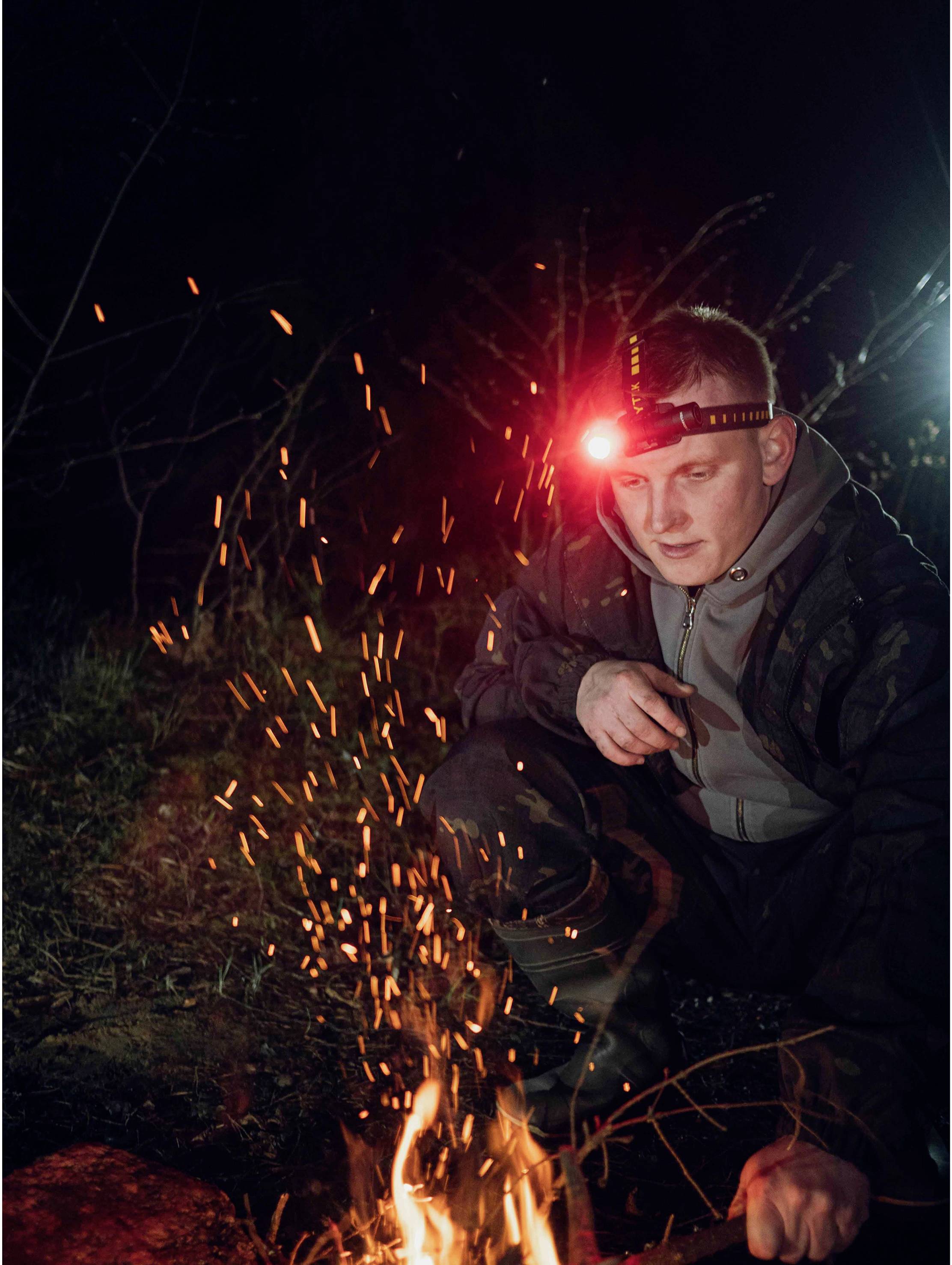 A person kneels outdoors at night beside a campfire, wearing a head torch that is illuminated. Sparks rise from the fire.
