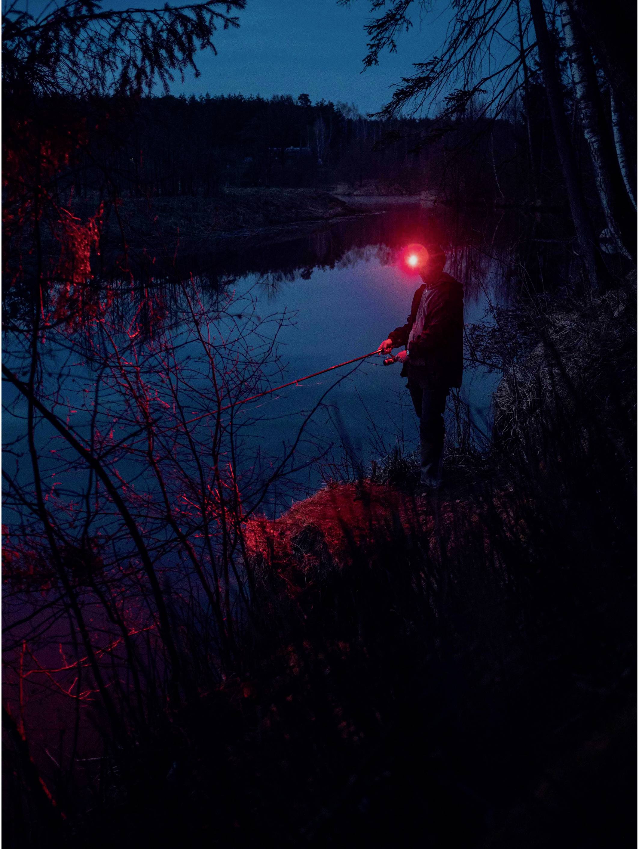 A person is fishing at night by a tranquil lake, surrounded by trees. A red light from their head torch reflects on the water's surface.