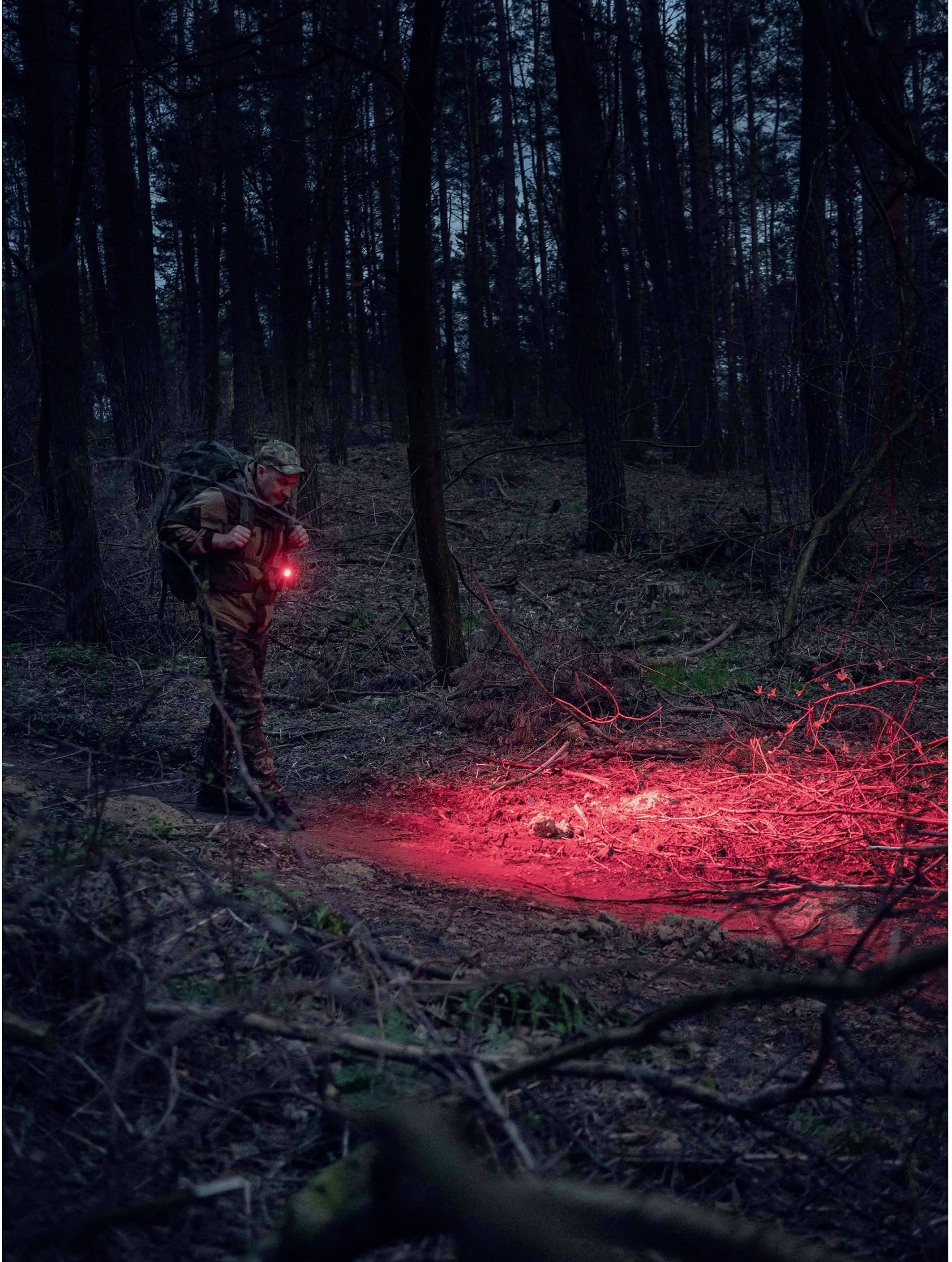 A person in the forest at dusk with a red light. The ground in front of them is also illuminated in red. The surroundings are densely wooded.