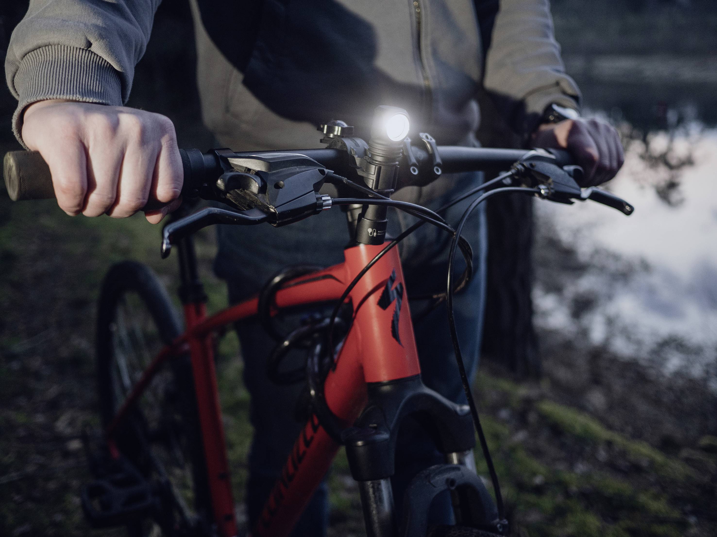 A person is holding a red bicycle by the handlebars; a beam of light shines from the front. The surroundings are wooded and twilight.