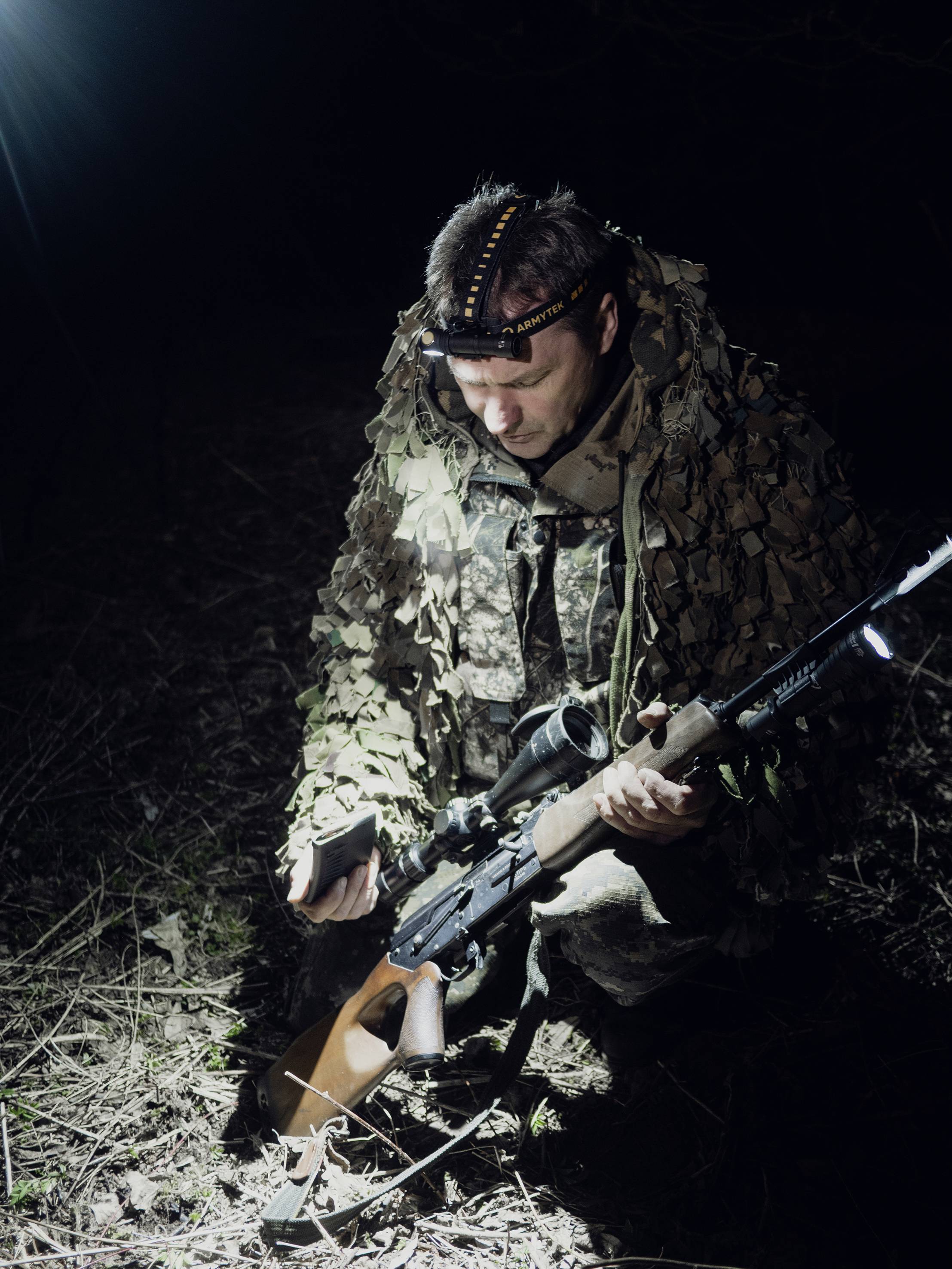 A person in camouflage clothing kneels on the ground in the darkness, holding a rifle. They are illuminated by an intense light.