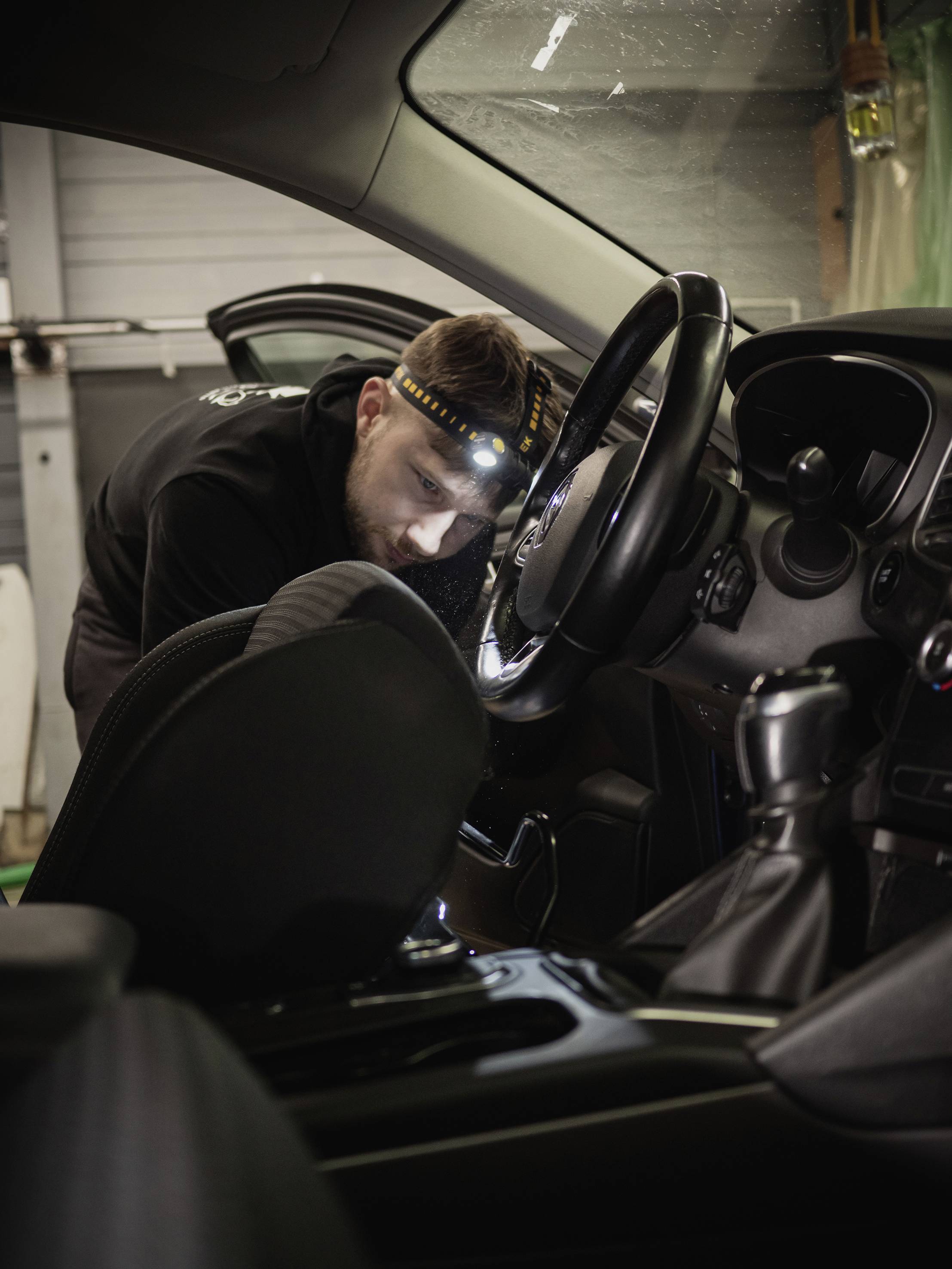 A mechanic is wearing a head torch and working inside a car, focused on the area around the steering wheel and dashboard.