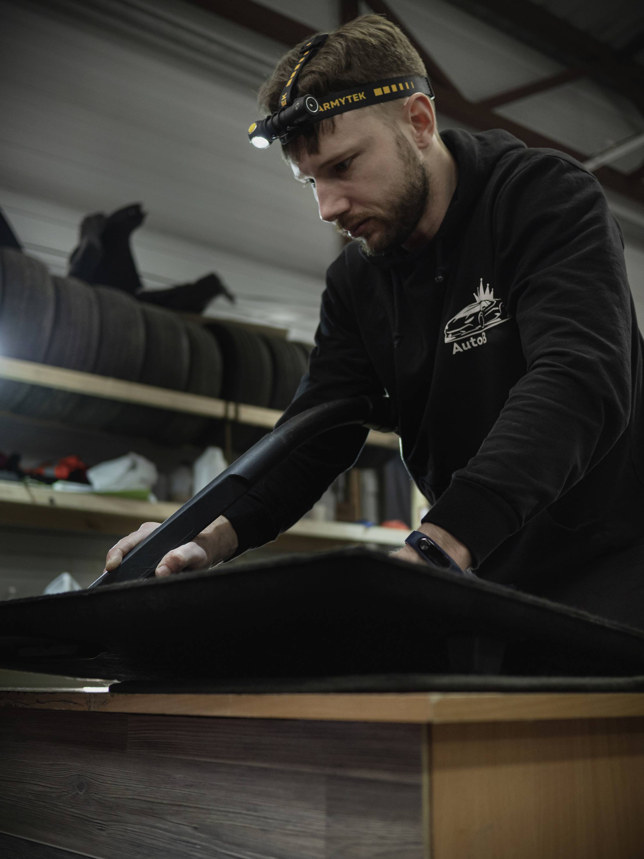 A man is working intently with a black cloth in a workshop, wearing a head torch and a black jumper.