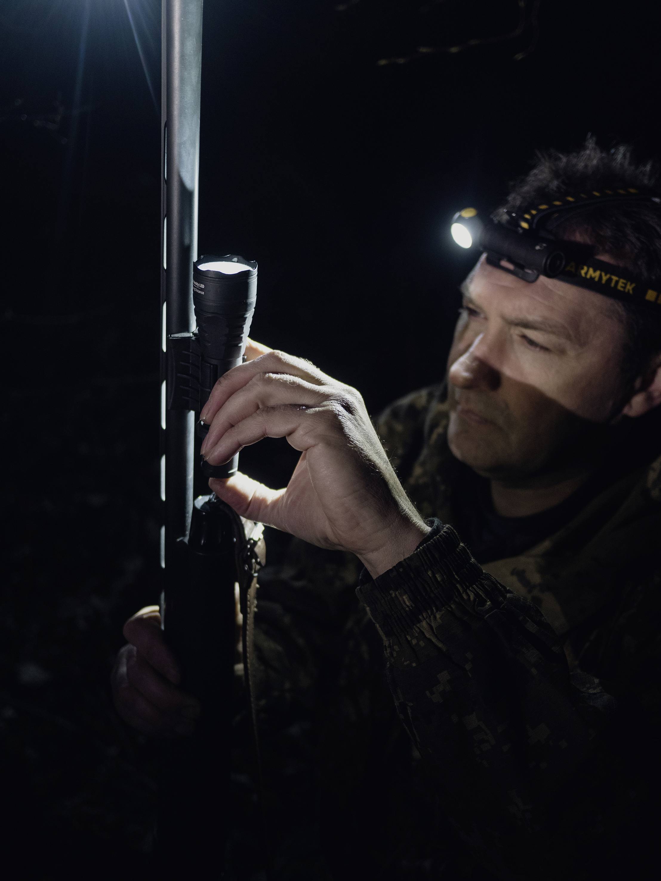A person wearing a headtorch is holding and examining a rifle or similar object in a dark environment.