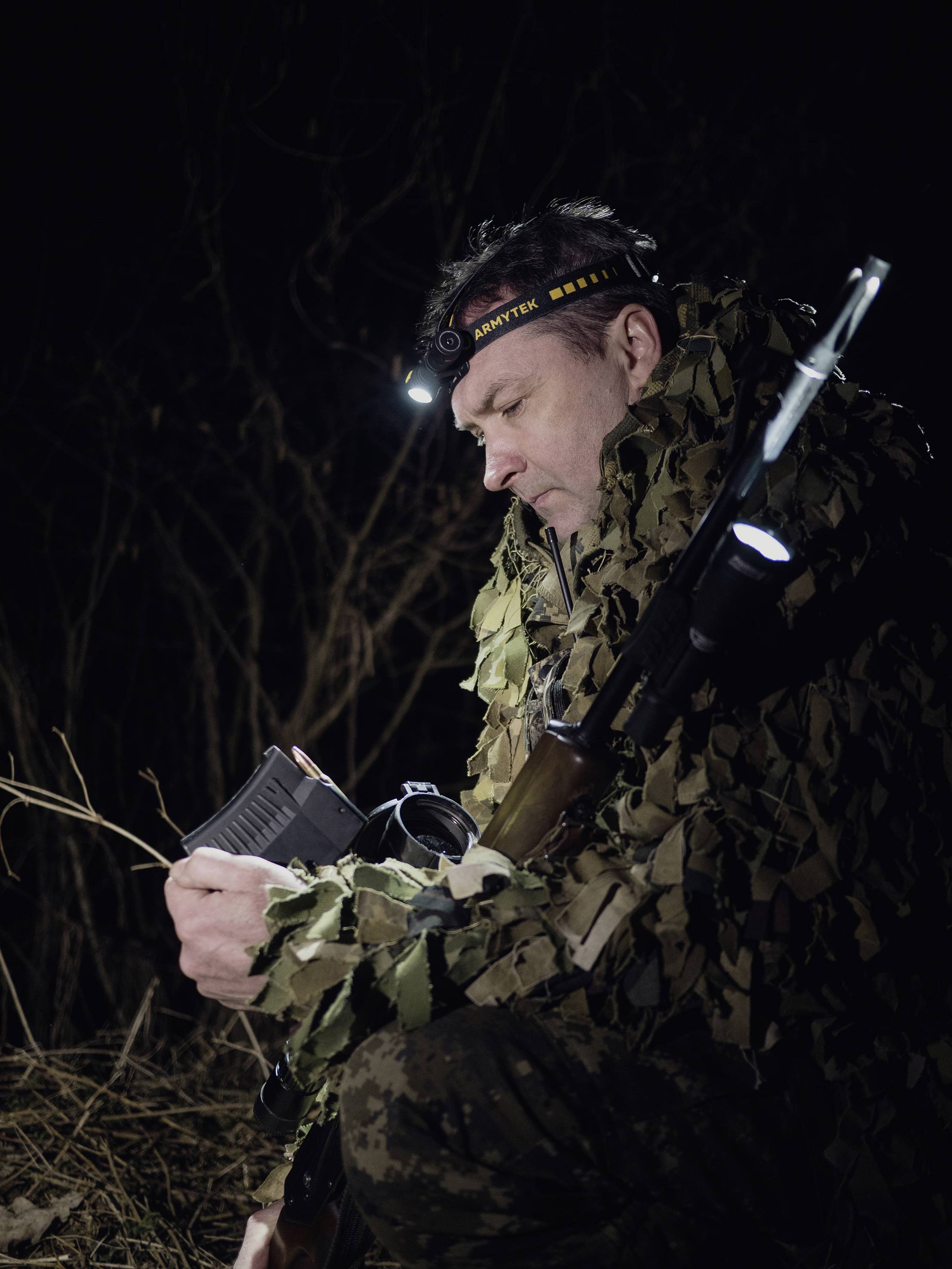 A person in camouflage clothing sits outdoors in the darkness, checking a magazine with a head torch. They are holding a rifle.