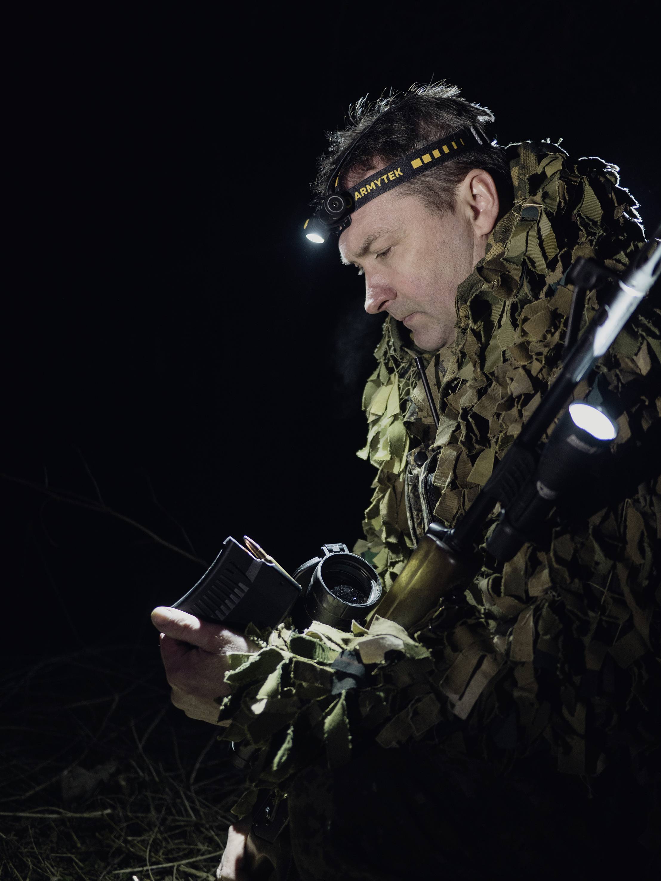 A person in camouflage gear is reading in a notebook using a head torch at night, while carrying a rifle.