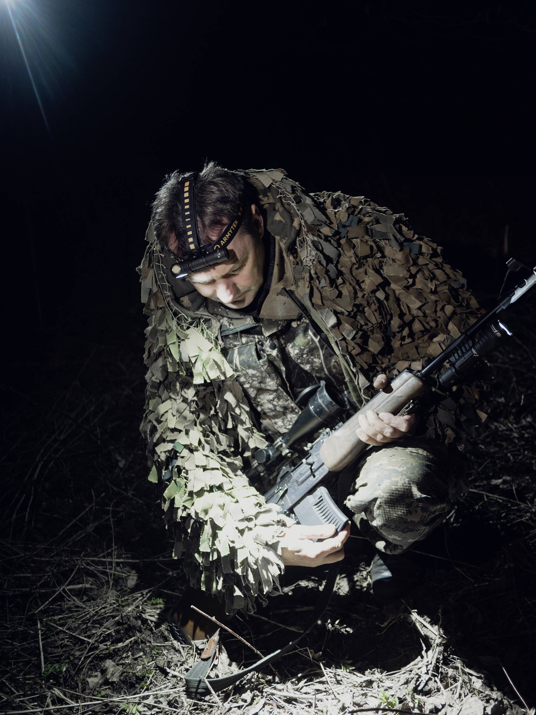 A soldier in camouflage gear kneels in a dark terrain, holding a rifle, illuminated by a powerful beam of light.