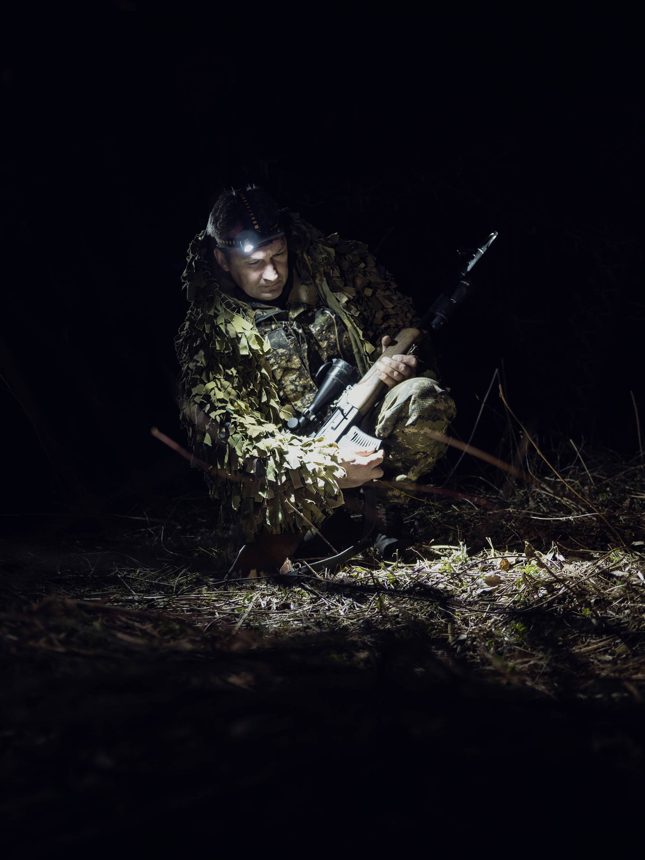 A person in camouflage kneels at night in the woods and holds a rifle. A spotlight illuminates the scene as they gaze at the ground.
