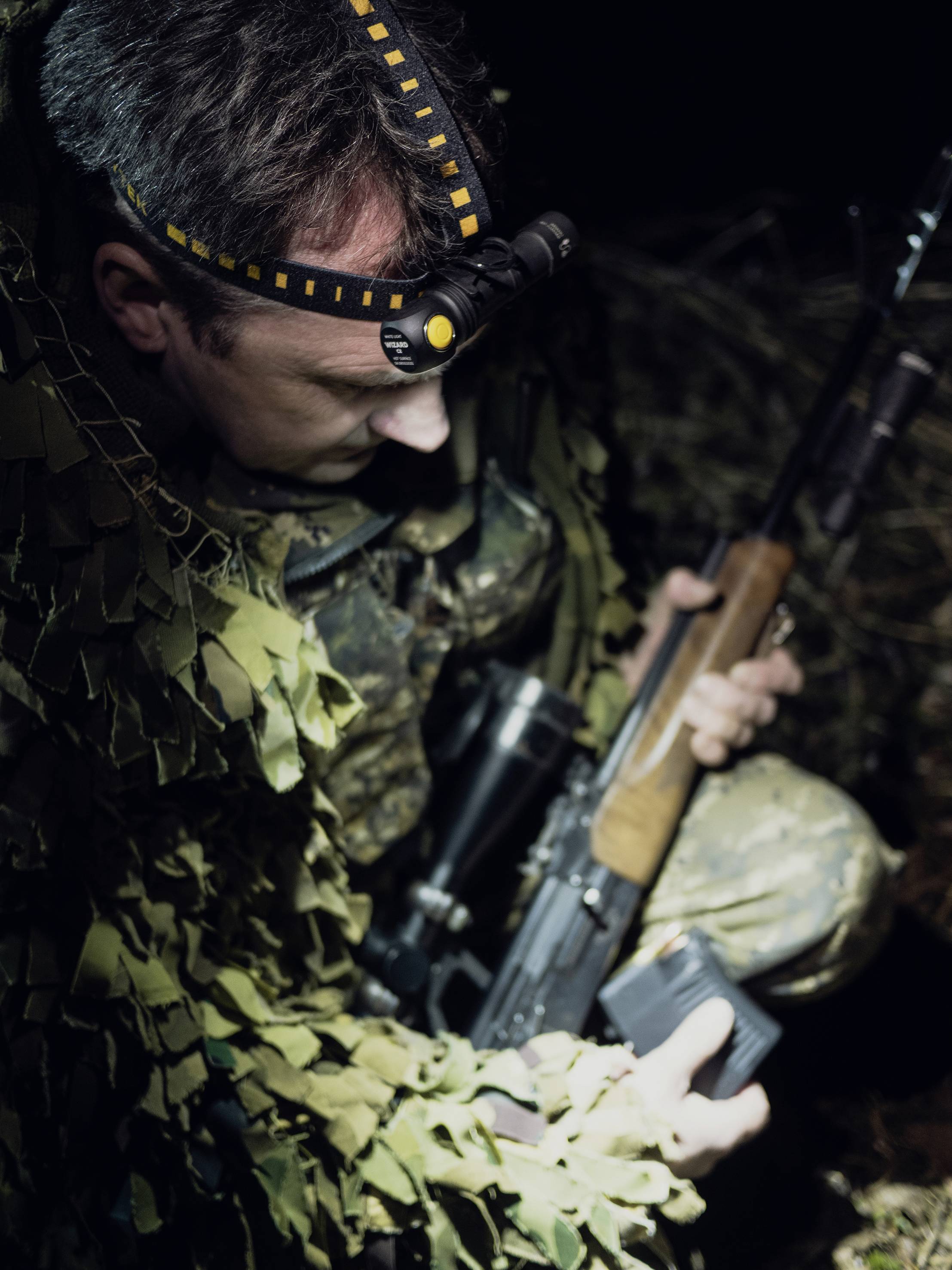 A person in camouflage clothing is holding a rifle and wearing a head torch. They appear to be in a dimly lit woodland area.