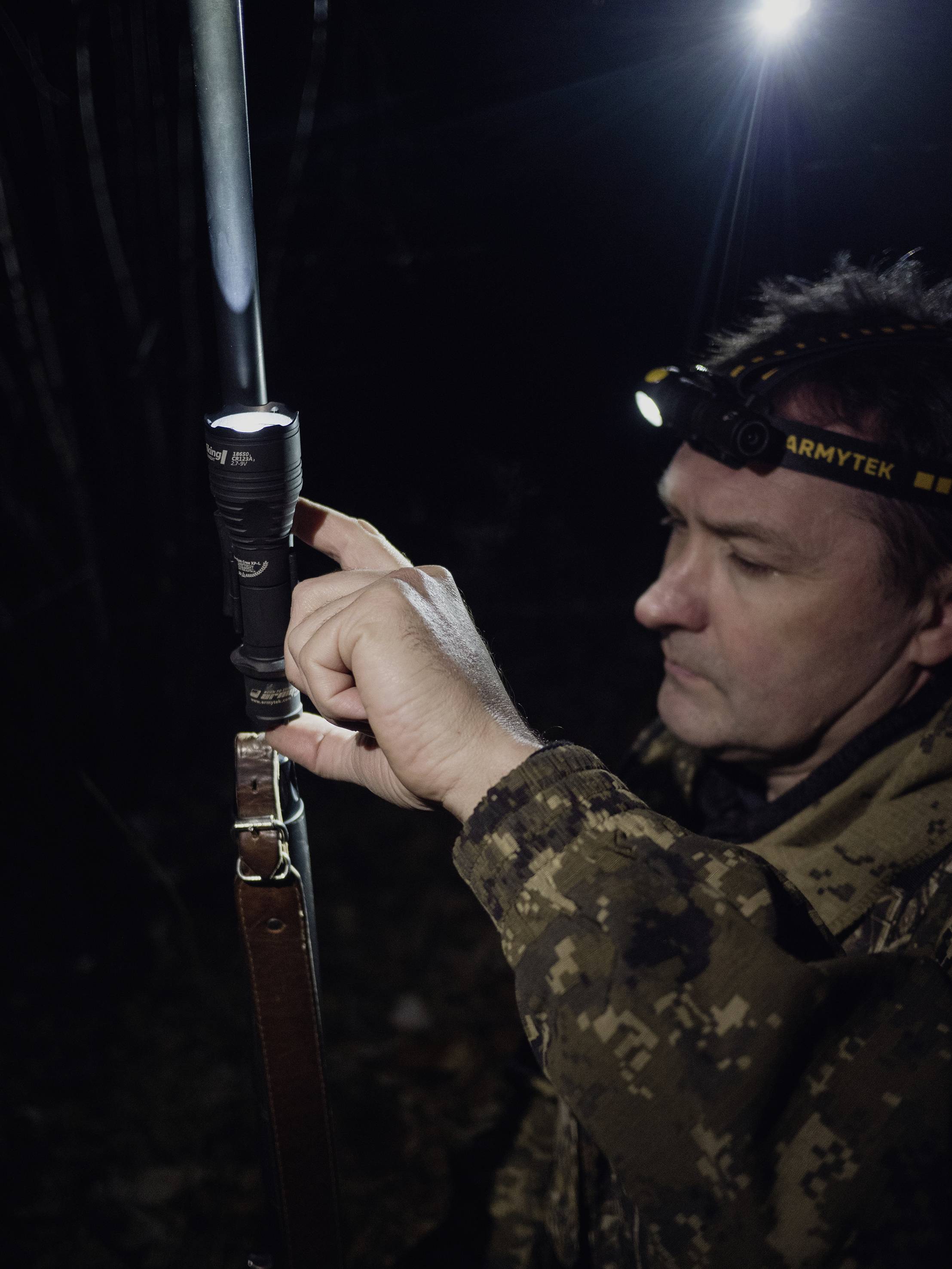 A person in camouflage gear adjusts a clipped torch in the darkness. They are wearing a head torch and standing in a wooded area.