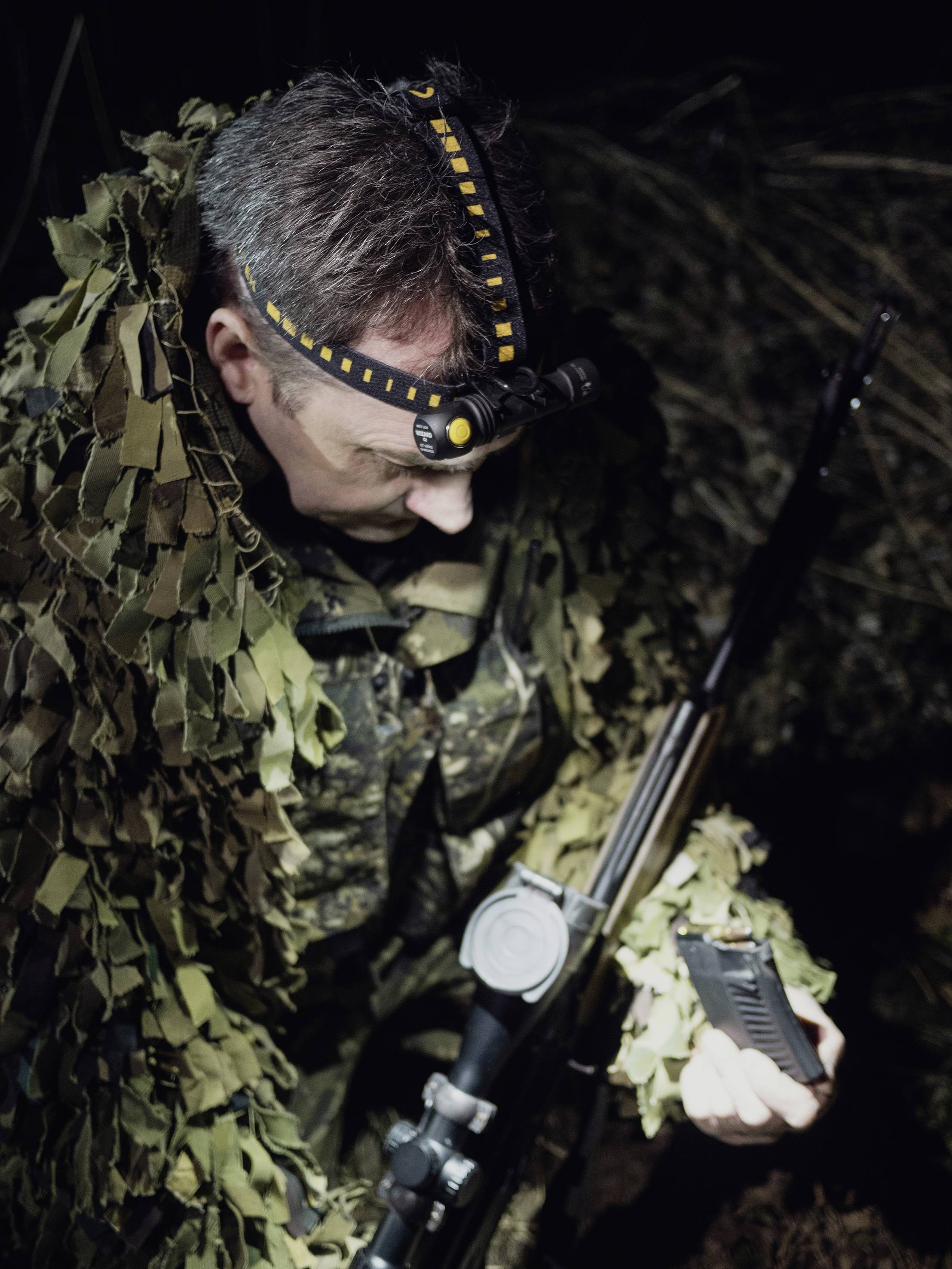 A person in camouflage clothing is holding a rifle and a magazine, presumably during a nocturnal hunt or military exercise.