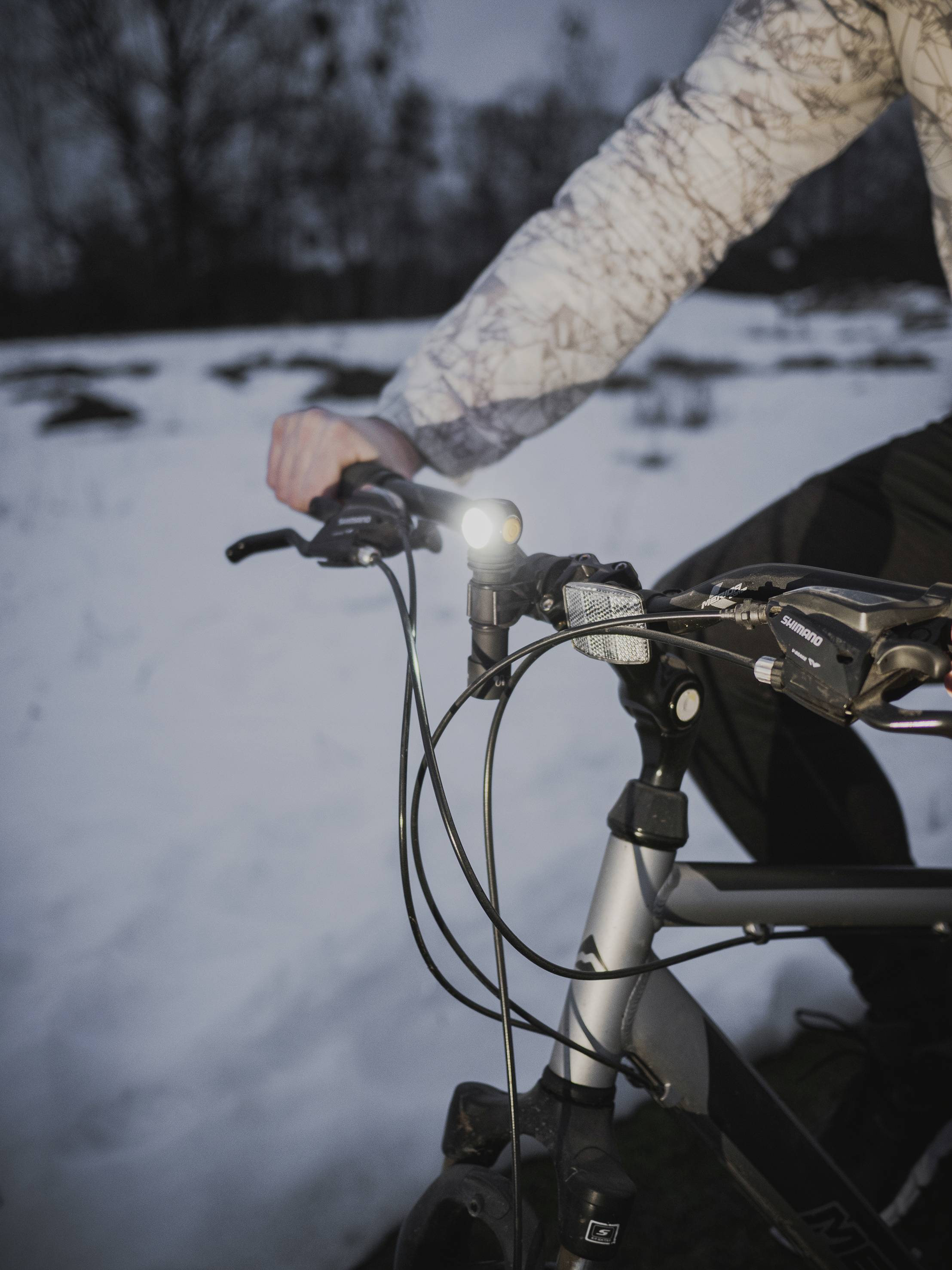 A person is cycling through the snow, wearing a patterned jacket. The bicycle light is illuminated, indicating it is either evening or early morning.