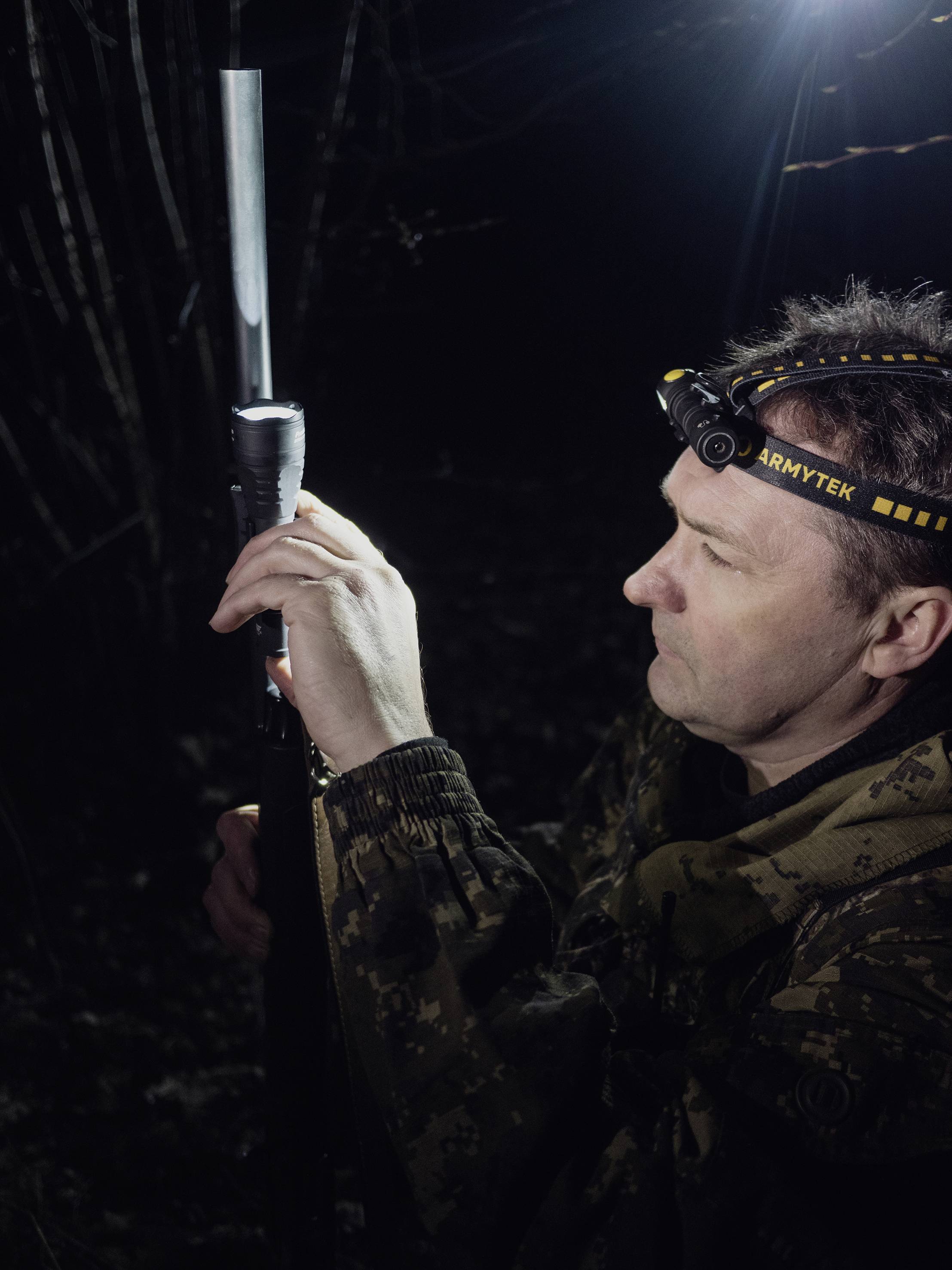 Man in a military-style suit in the darkness. He is holding a long piece of equipment and wearing a head torch.