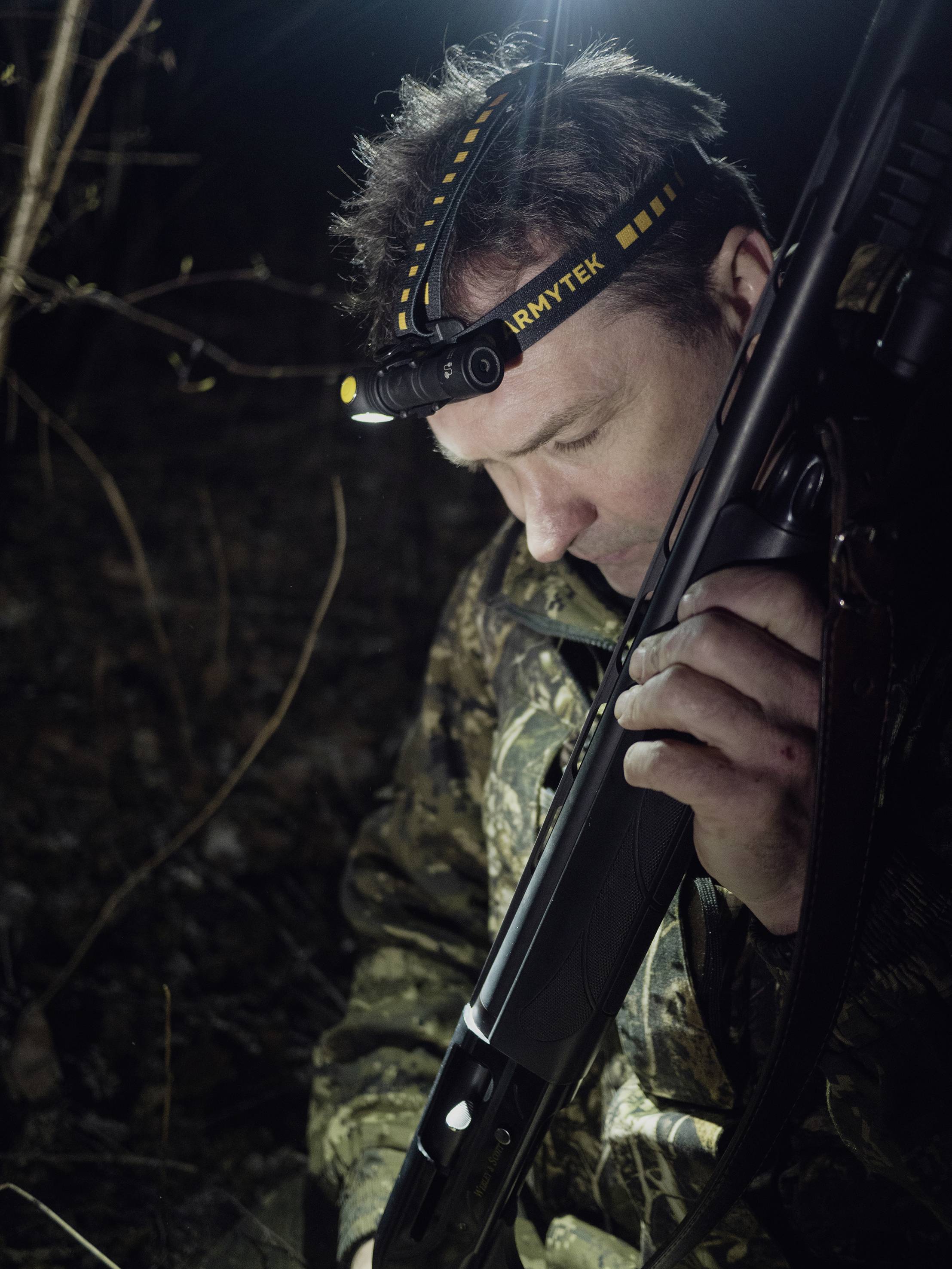 A man in camouflage clothing is holding a rifle and wearing a head torch while standing in a dark forest.