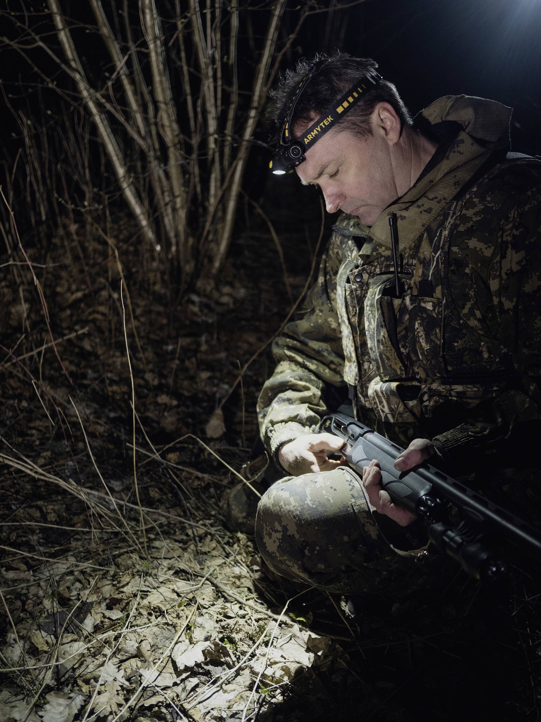 A man in camouflage kneels in the forest at night, holding a rifle. A head torch illuminates the ground and some dry branches.