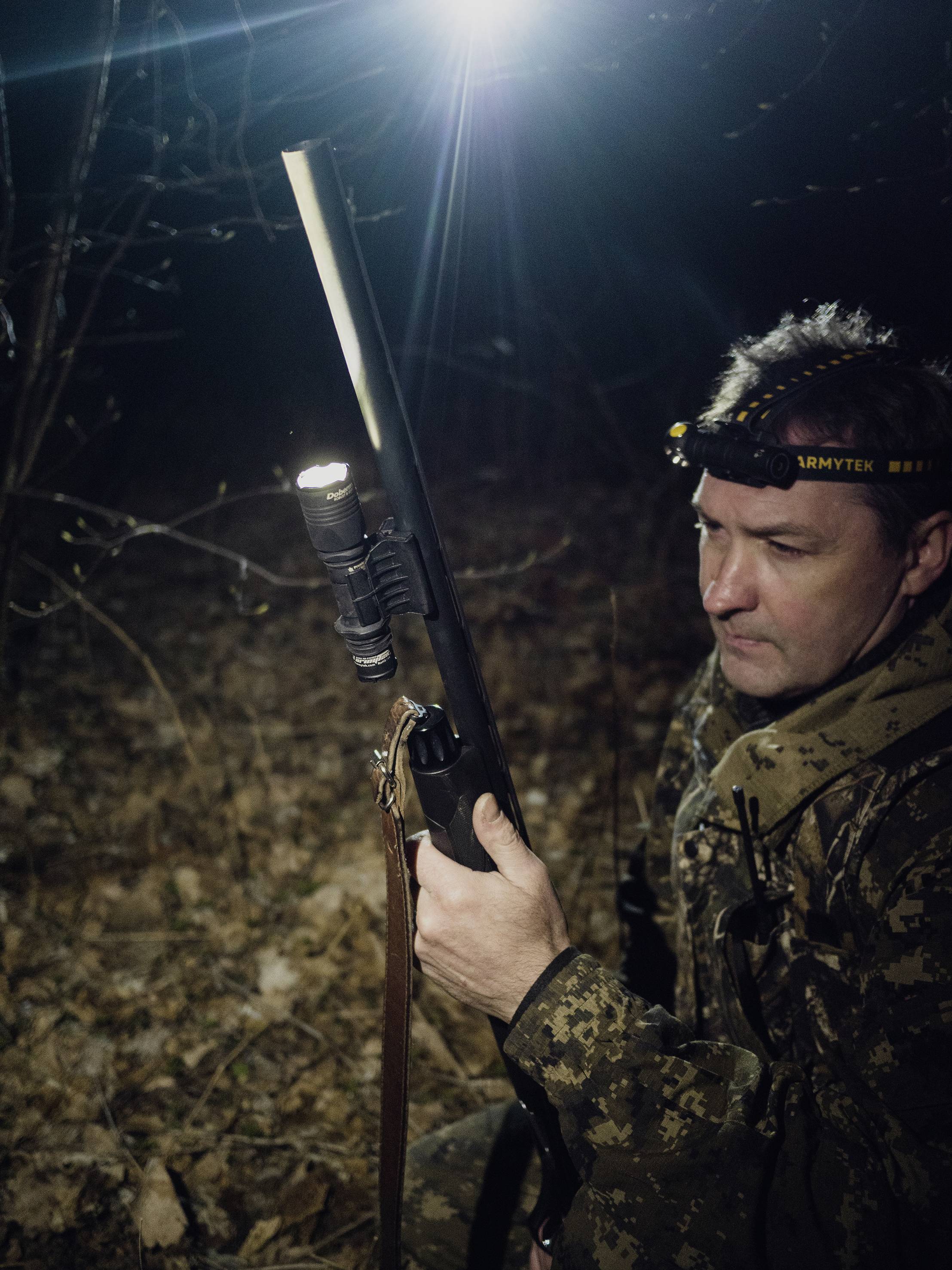 A person in camouflage clothing holds a hunting rifle with a torch at night, surrounded by woodland. A head torch illuminates the path.