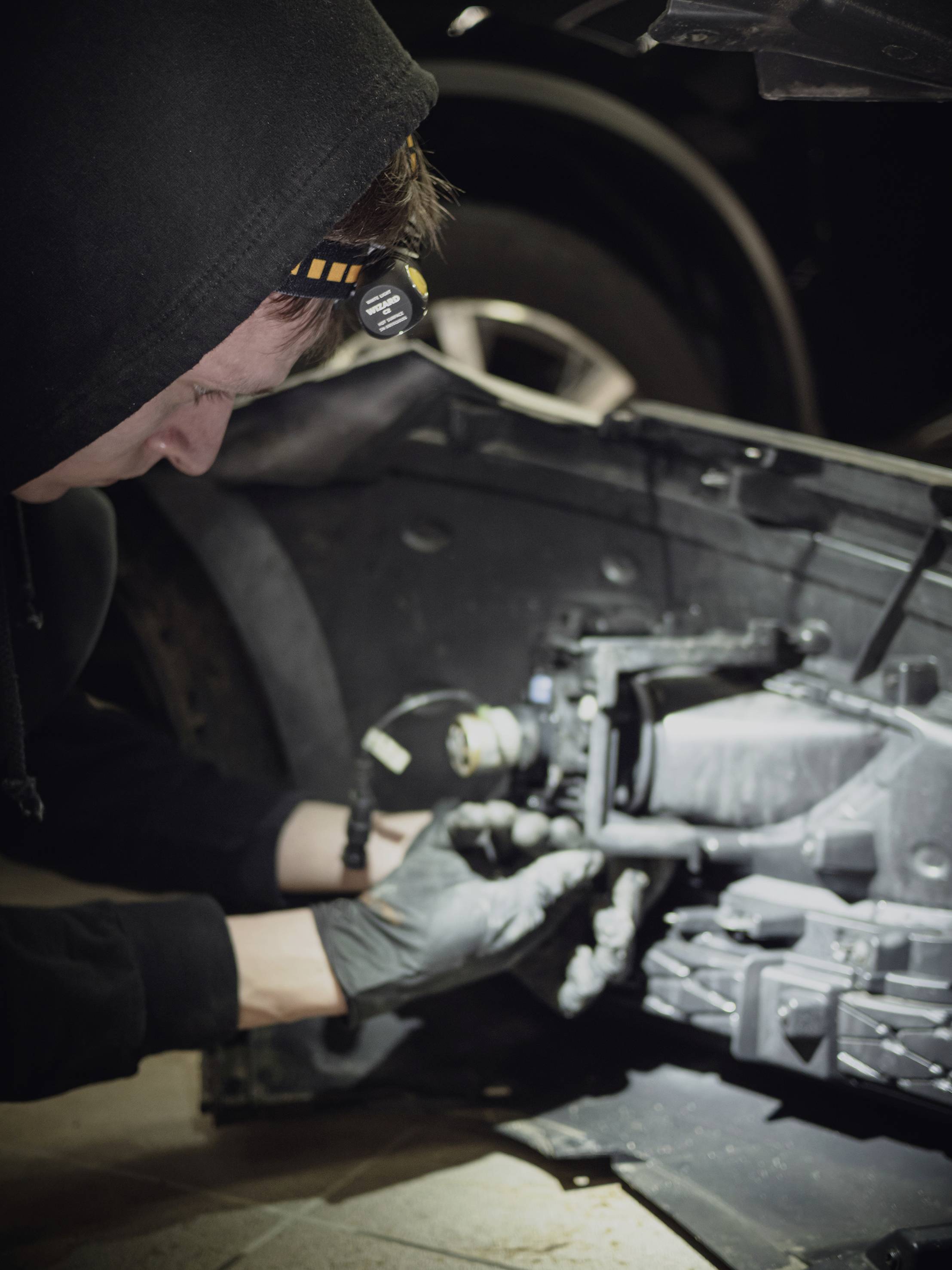 A mechanic in dark clothing and wearing a head torch is repairing a car part in a workshop. A tyre is visible in the background.