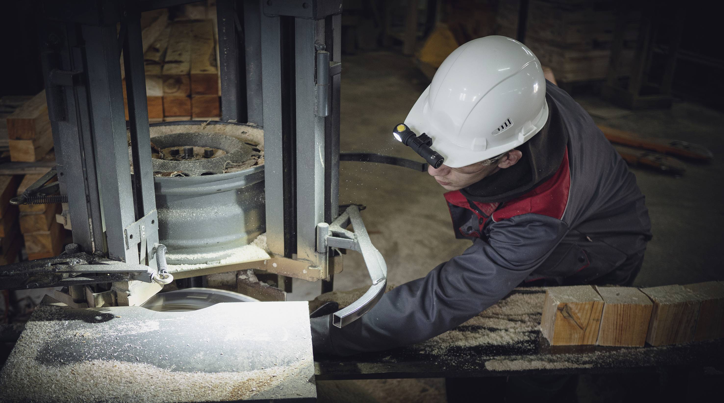 A worker in protective clothing operates a woodworking machine in a workshop. Wood shavings and wood pieces are visible on the table.