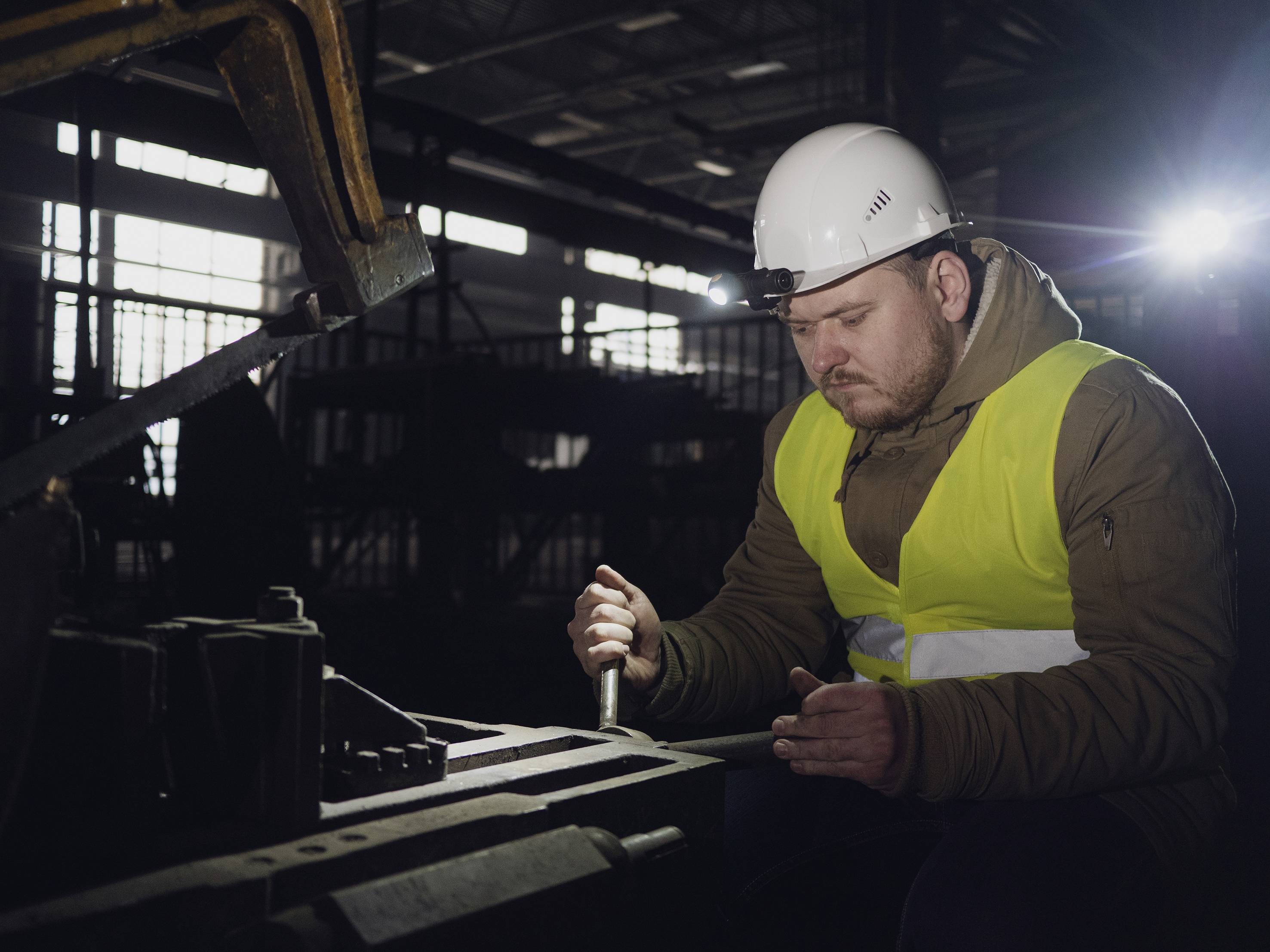 A worker in safety clothing is working on a metal piece with a tool in a factory hall; subdued lighting in the background.