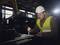 A worker in safety clothing is working on a metal piece with a tool in a factory hall; subdued lighting in the background.
