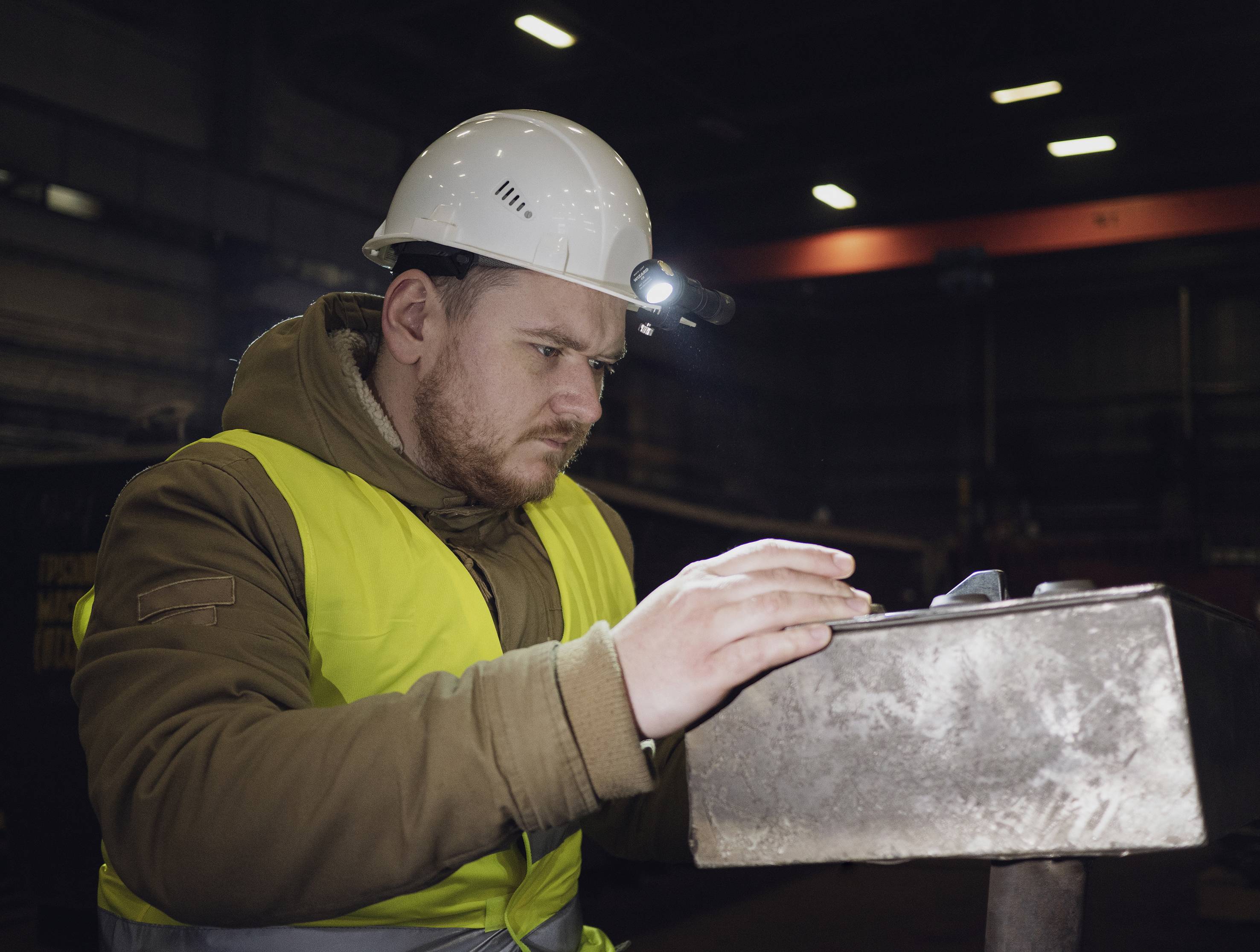 A man wearing a yellow high-visibility vest and hard hat working in a factory hall. He is concentrating intently on a metal component.