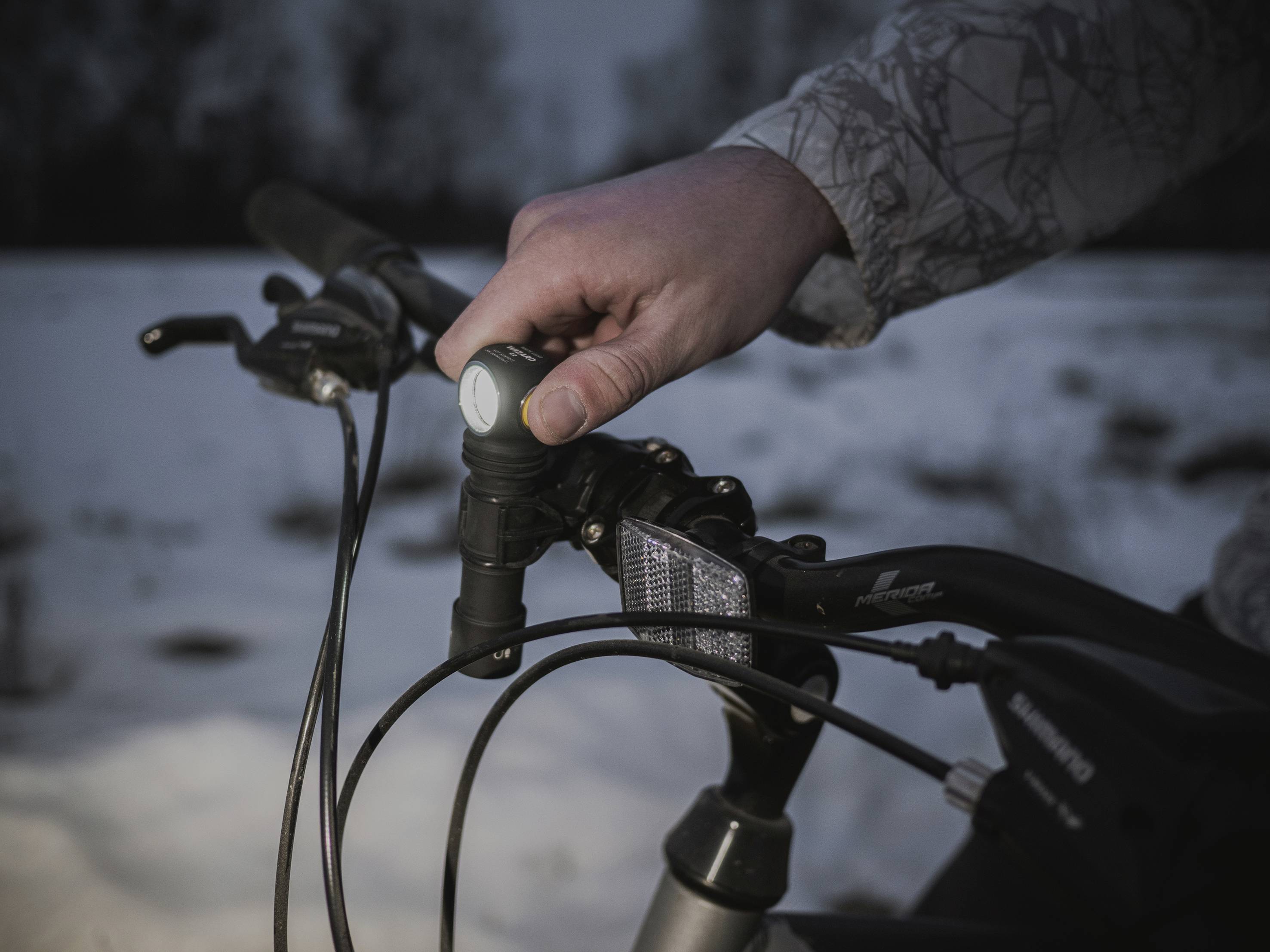 A person switches on the front light of a bicycle at dusk. The background is snow-covered.
