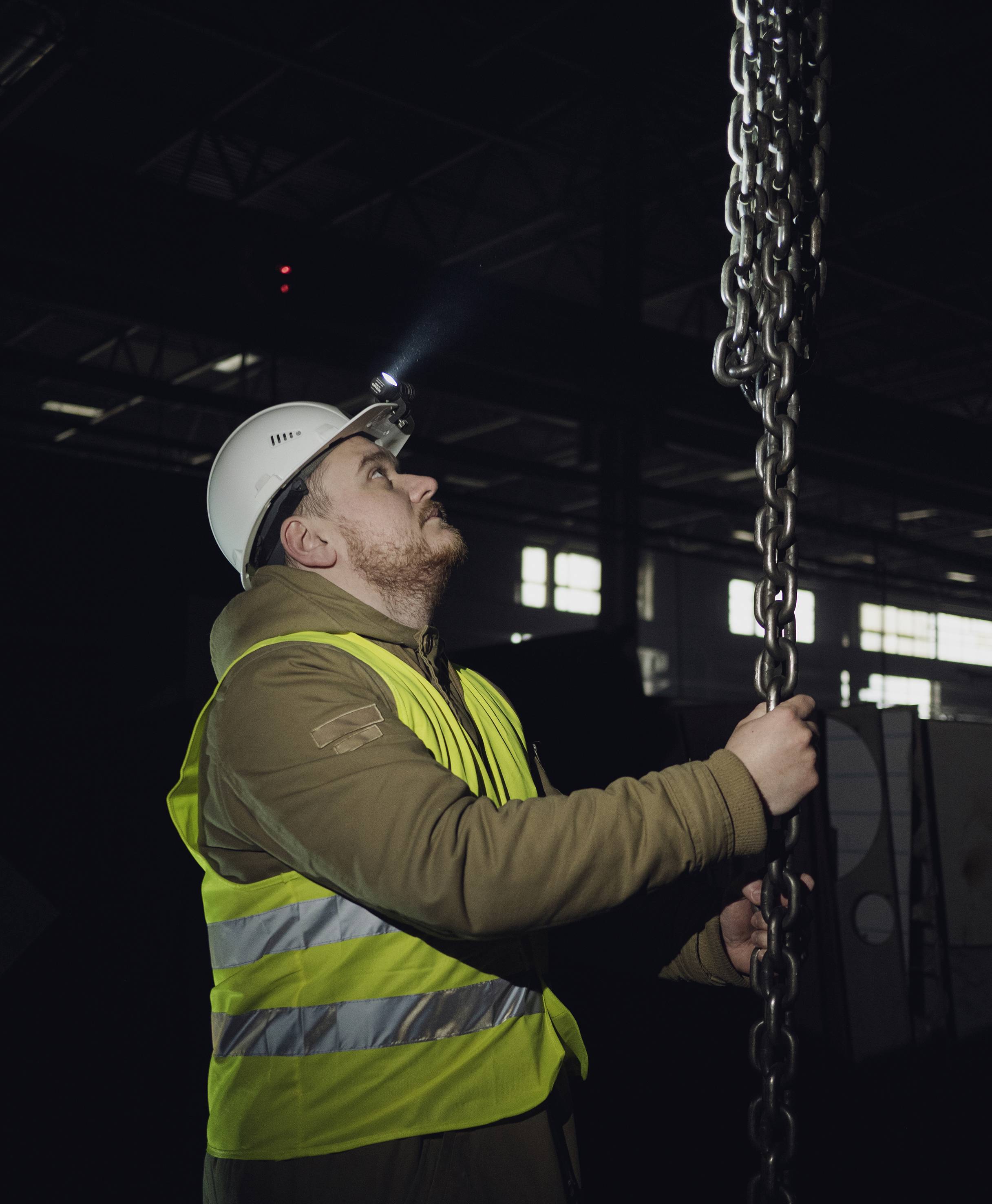 A worker wearing a hard hat and high-visibility vest is inspecting a heavy chain in a factory hall with dim lighting.
