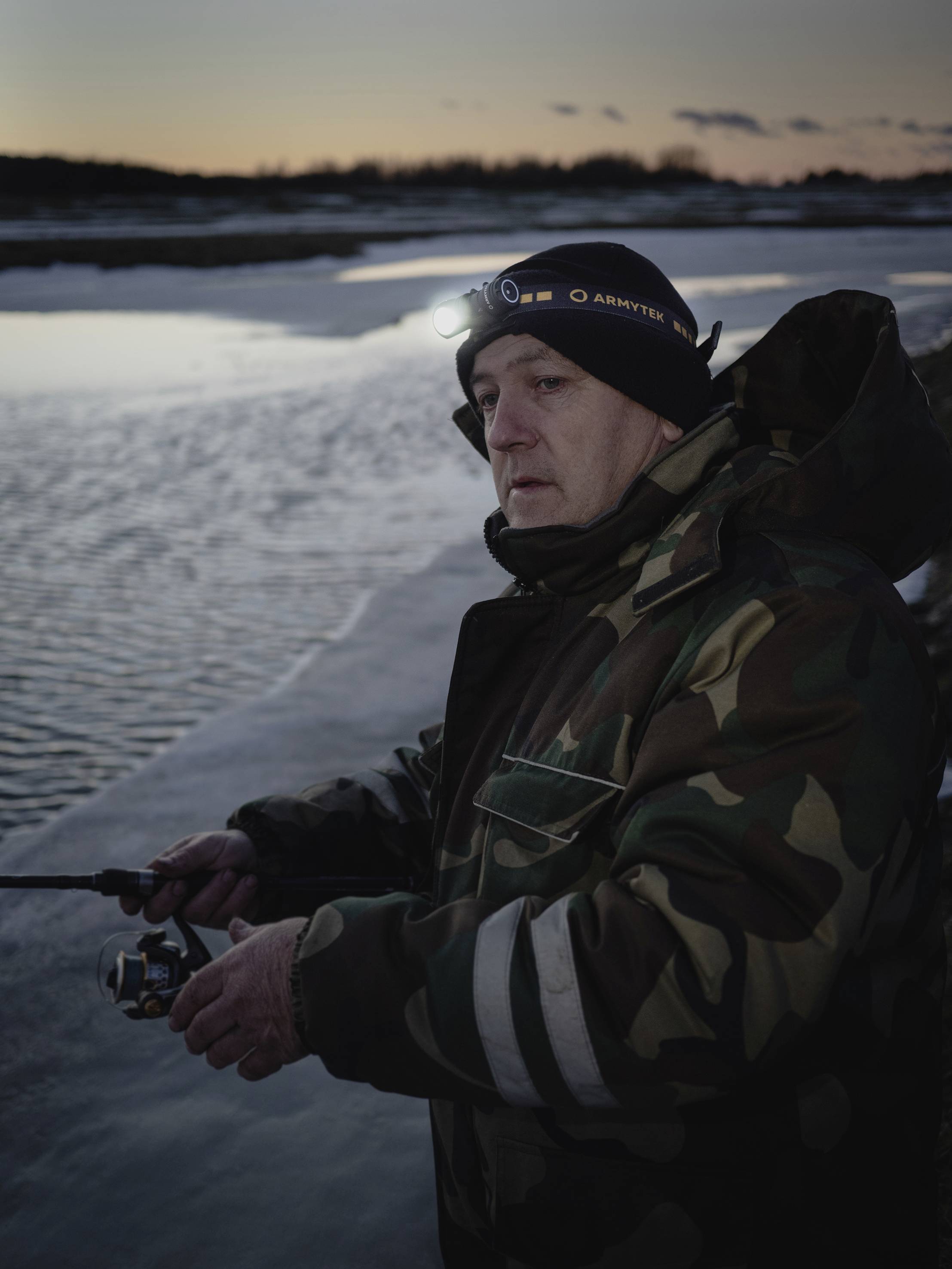 A man is fishing by a river at sunset. He is wearing a winter jacket and a beanie with a head torch.
