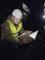 A construction worker in a yellow hi-vis vest and hard hat is examining an open book or document in a dimly lit room.