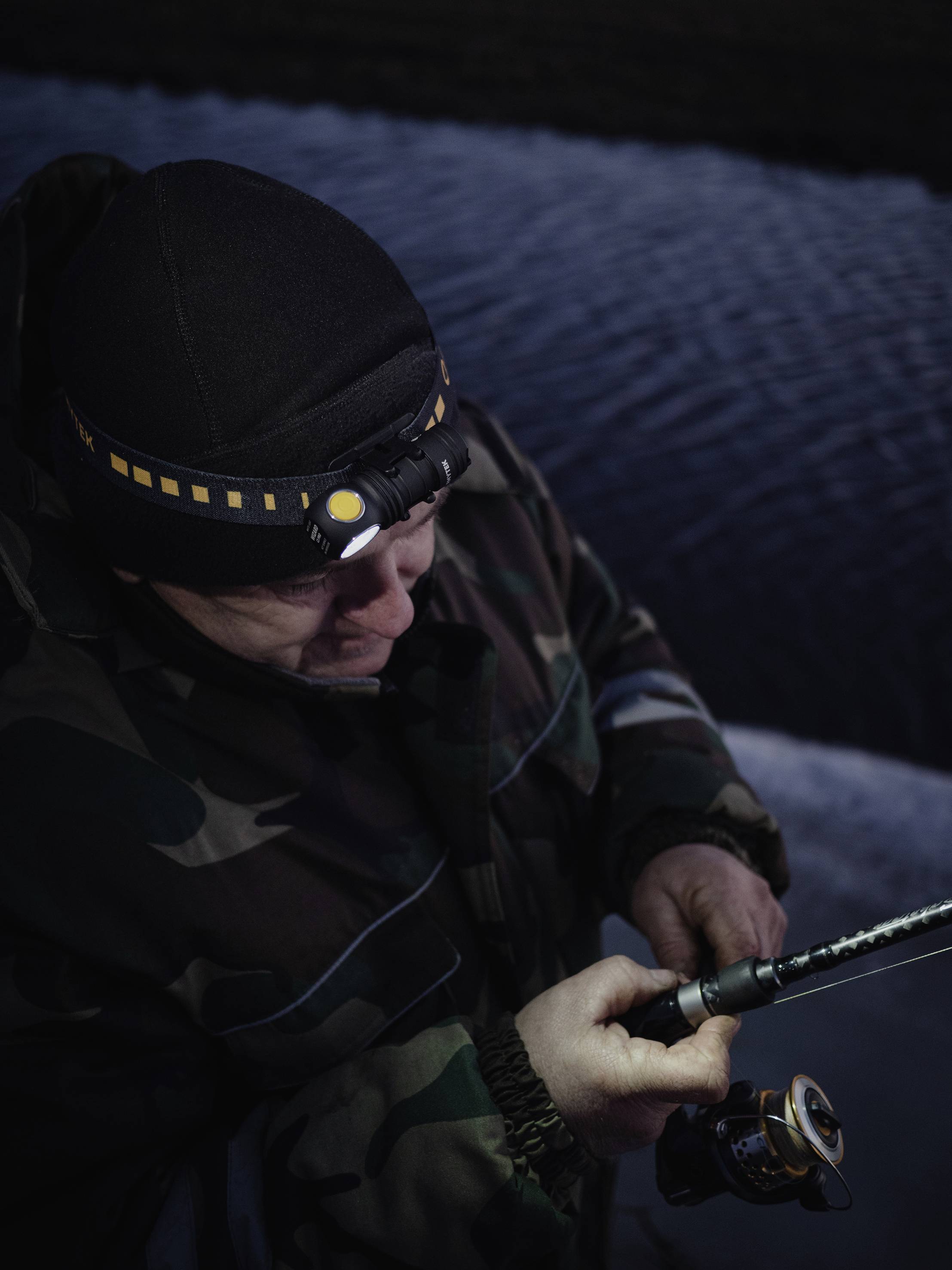 A person in camouflage clothing is fishing at night by the water, wearing a head torch.