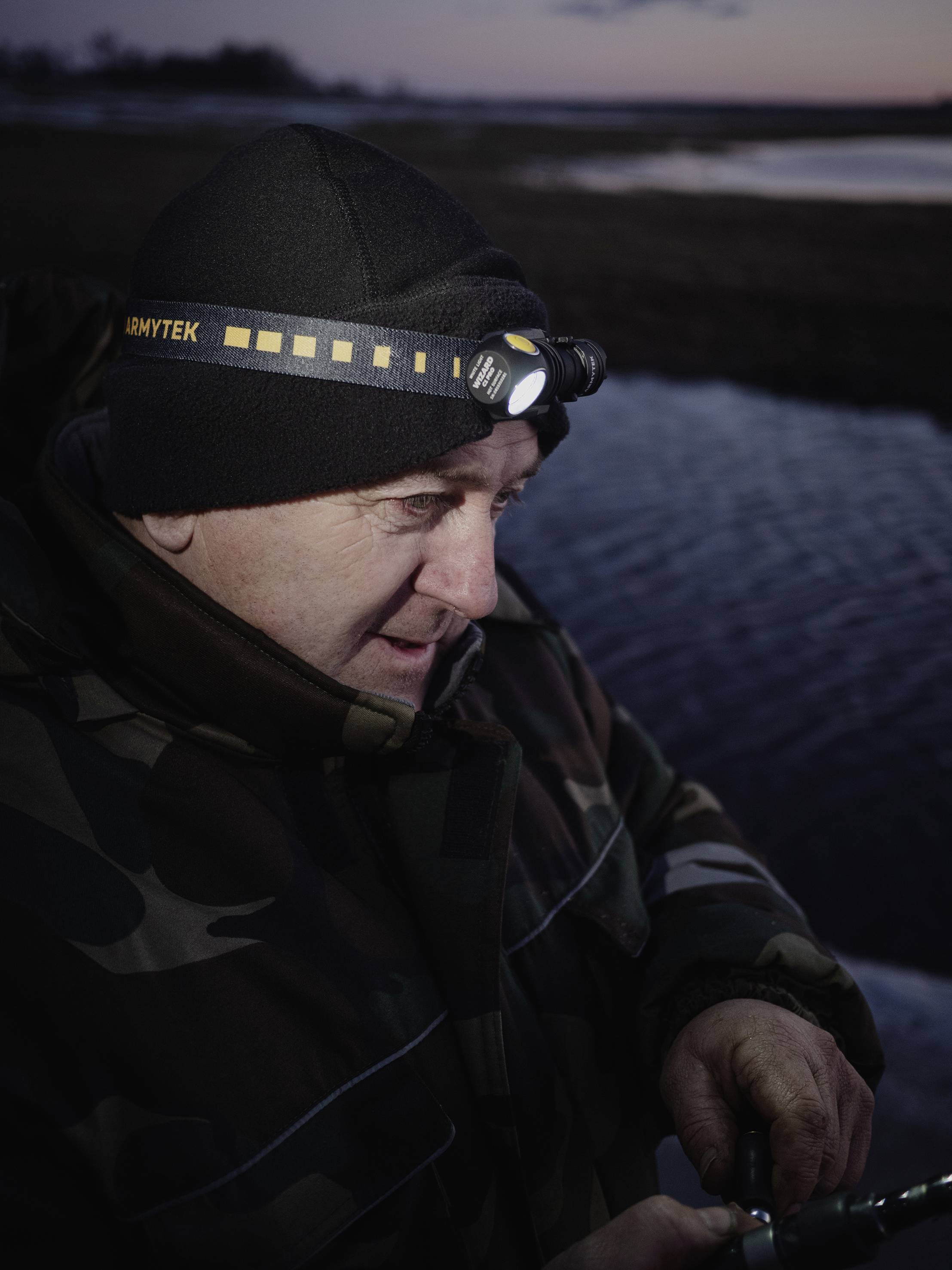 A person wearing a head torch fishes outdoors at dusk, wrapped up warmly in a camouflage jacket. The background shows water and a dark sky.
