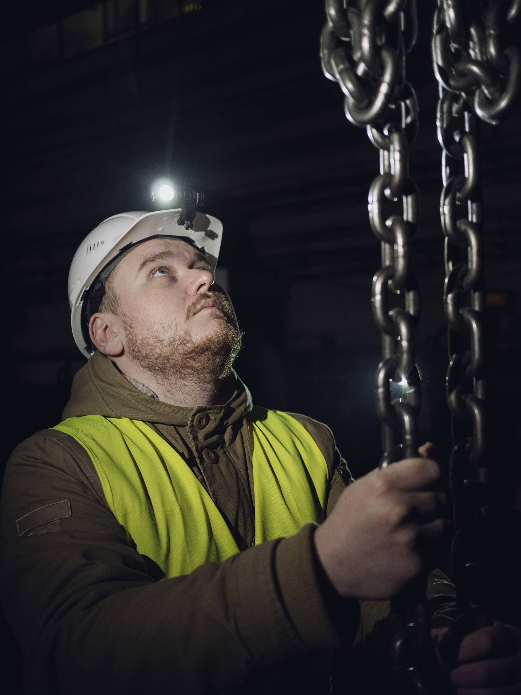A worker wearing a hard hat and high-visibility vest inspects heavy chains in a dimly lit environment, focused and looking upwards.