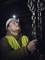 A worker wearing a hard hat and high-visibility vest inspects heavy chains in a dimly lit environment, focused and looking upwards.