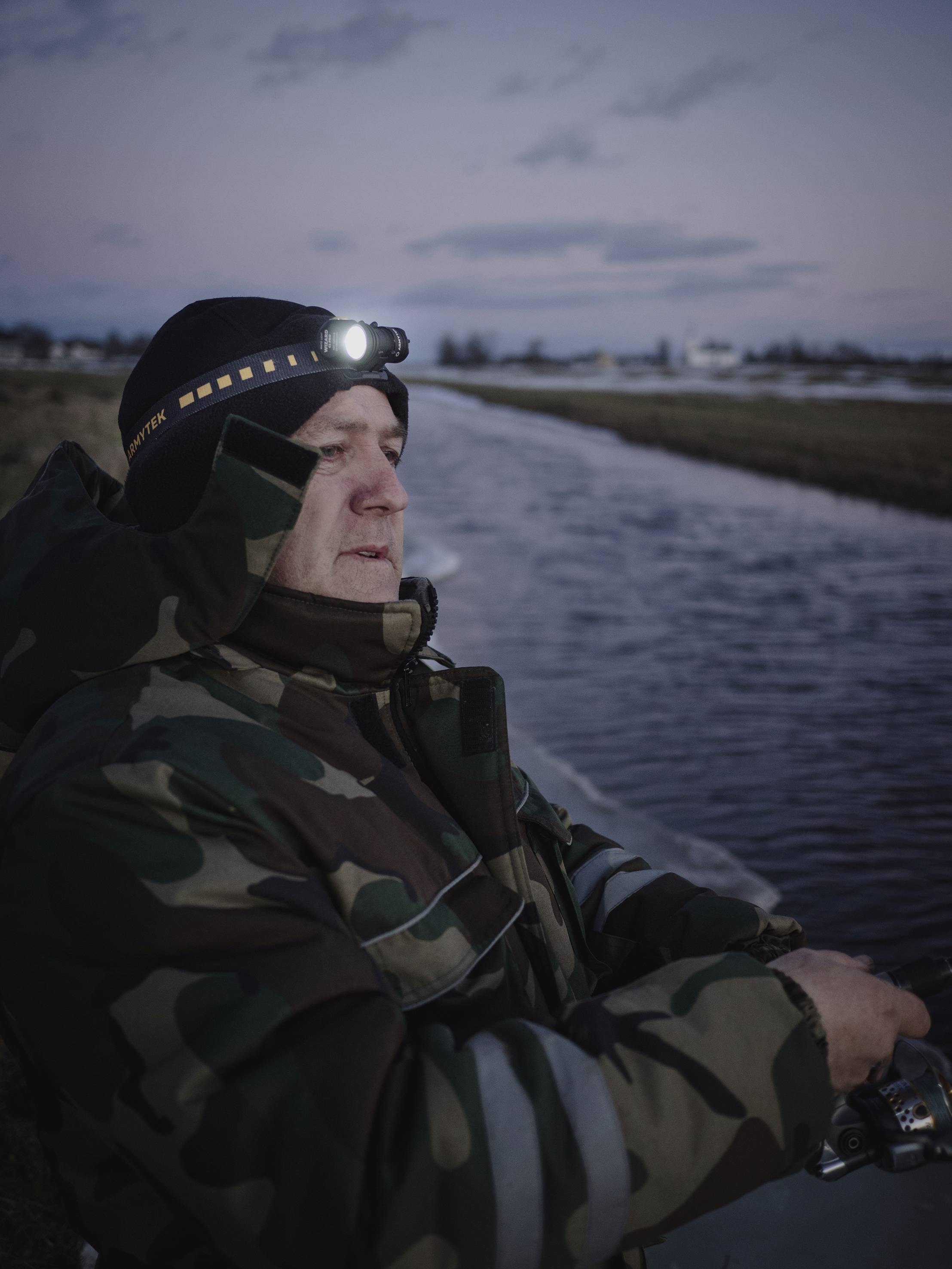 A fisherman in camouflage clothing stands on the riverbank at dusk, wearing a head torch. A flat landscape is visible in the background.